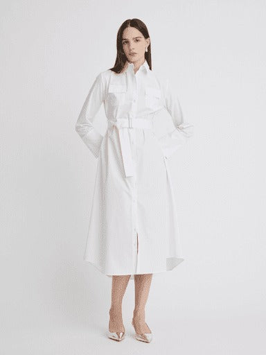 A woman stands against a plain white background, wearing the Matthew Bruch Cargo Button Up Midi White Poplin Shirt Dress. This long-sleeved, collared dress made of 100% cotton falls below her knees and features a belt at the waist. Made in the USA, she pairs it with white pointed-toe heels and has shoulder-length dark hair. Her hands rest casually in the dress pockets.