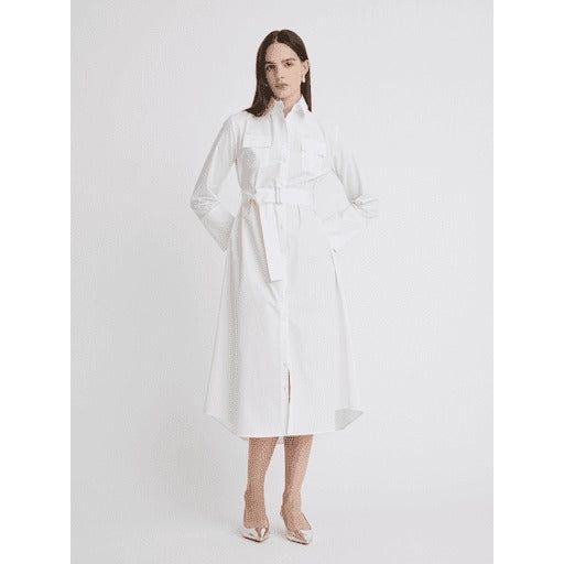 A woman stands against a plain white background, wearing the Matthew Bruch Cargo Button Up Midi White Poplin Shirt Dress. This long-sleeved, collared dress made of 100% cotton falls below her knees and features a belt at the waist. Made in the USA, she pairs it with white pointed-toe heels and has shoulder-length dark hair. Her hands rest casually in the dress pockets.