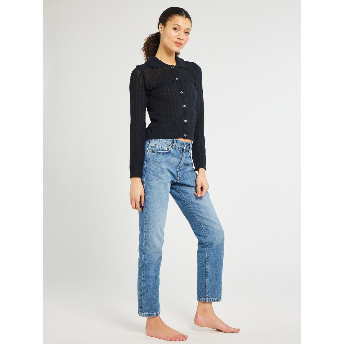 A woman stands on a plain background, wearing the black Mille Andie Cardigan with hand crochet trim and blue jeans. She is barefoot, with her right hand relaxed by her side and her left hand resting on her hip. She is smiling softly at the camera.