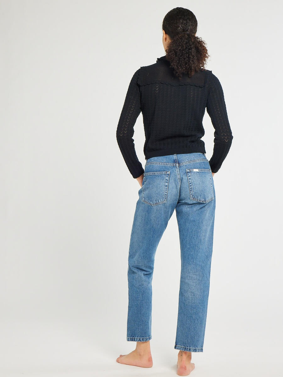 A person with curly hair is standing barefoot, facing away from the camera. They are wearing blue jeans and the Mille Andie Cardigan Black by Mille, featuring a hand crochet trim. The background is plain white.