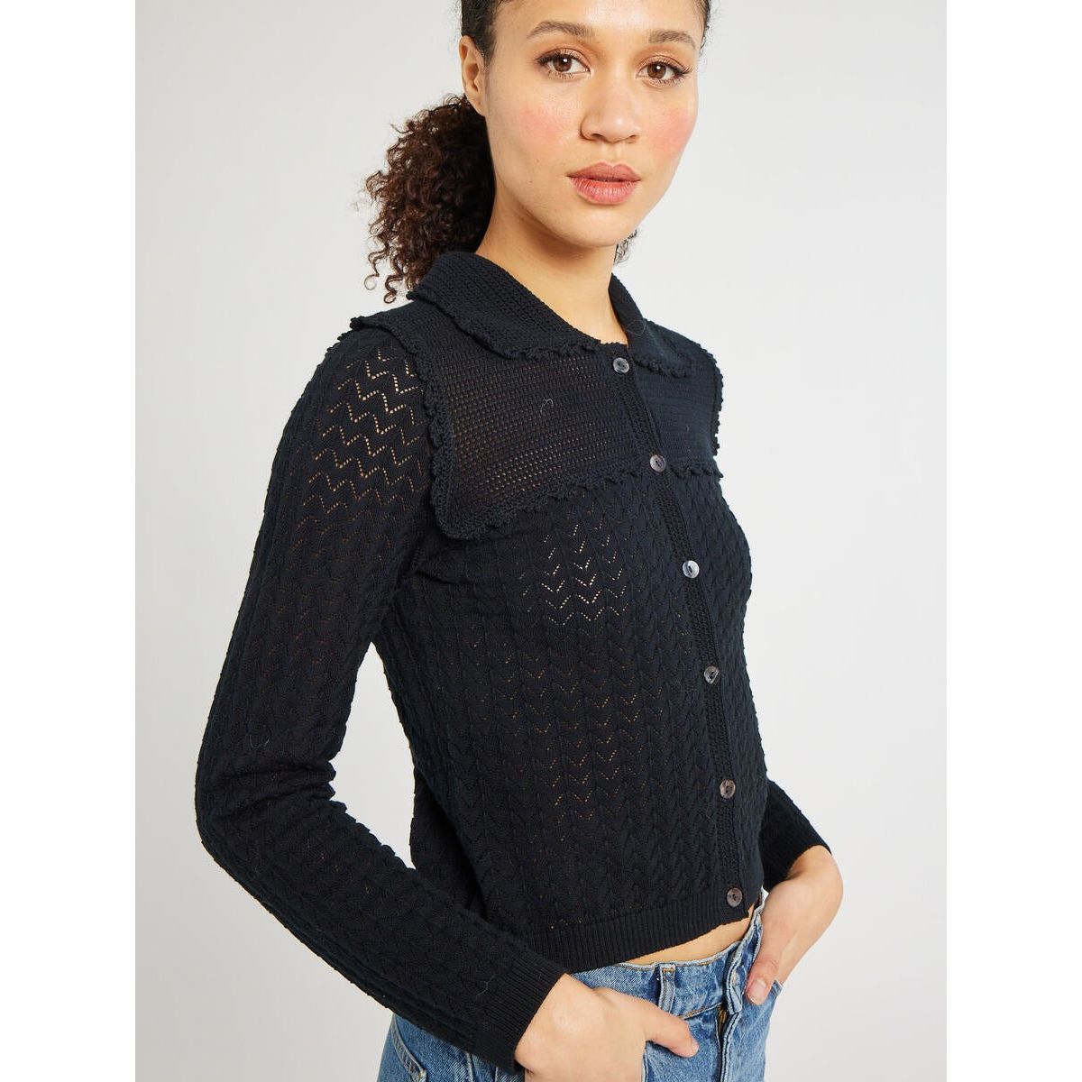 A person with curly hair stands against a plain background, wearing the Mille Andie Cardigan Black by Mille. The black, long-sleeved knit cardigan features buttons and intricate patterns, along with a delicate hand crochet trim. They have their hand in the pocket of their blue jeans and are looking directly at the camera.