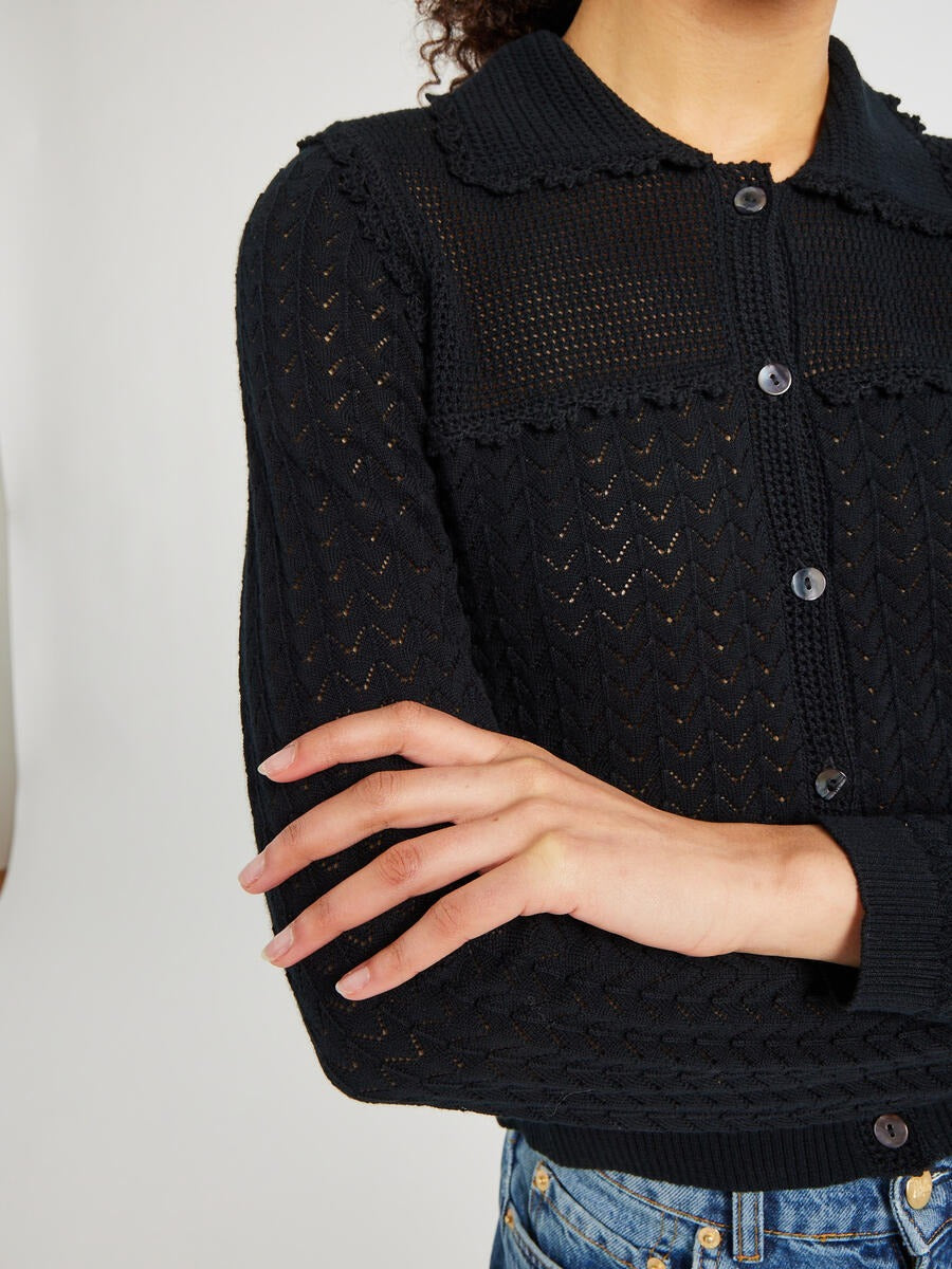 A person wearing the Mille Andie Cardigan Black by Mille, a long-sleeved, lightweight knit cardigan with a button-up front and a collar. They are crossing their arms and are shot from the shoulders down. The visible portion of their bottoms is a pair of blue jeans.