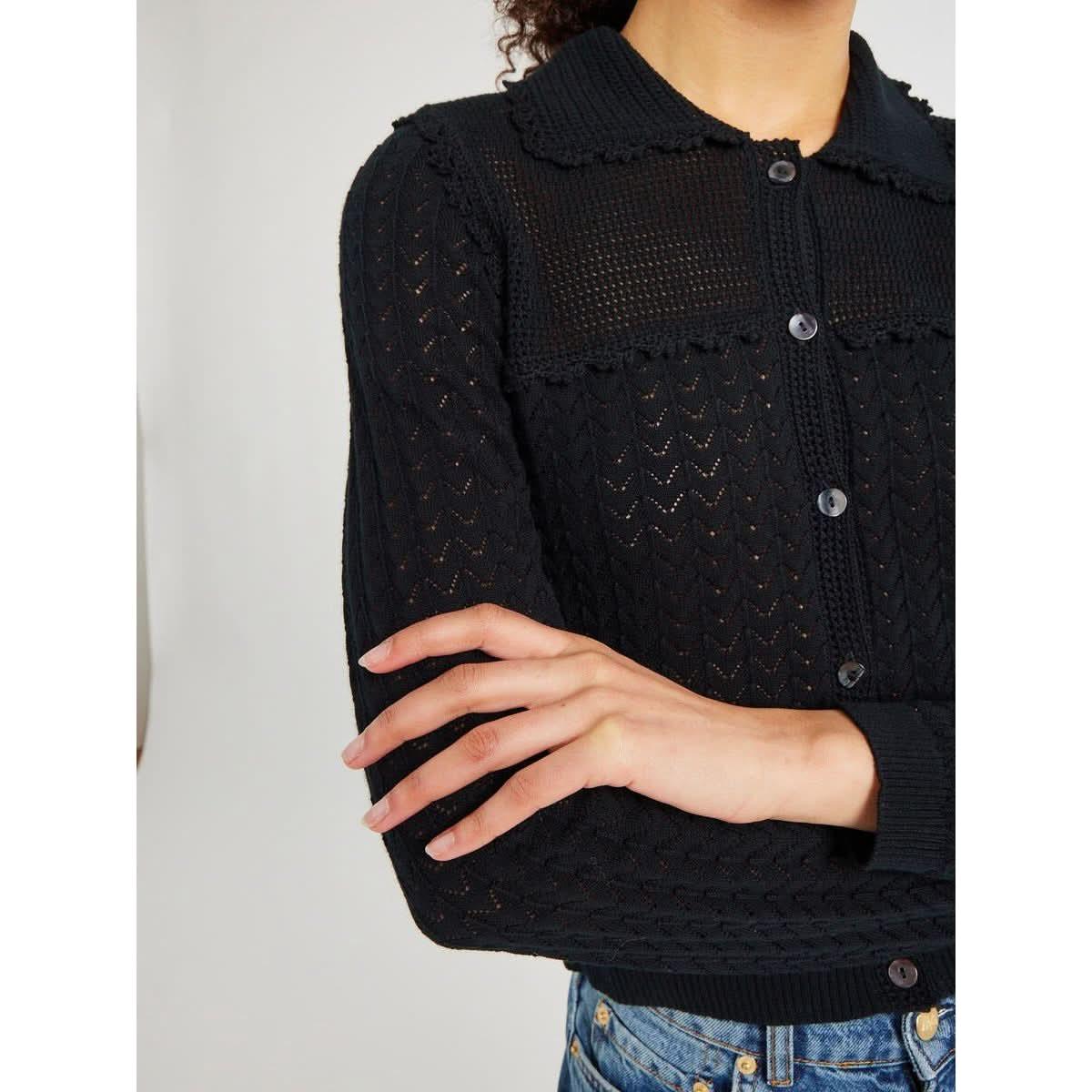 A person wearing the Mille Andie Cardigan Black by Mille, a long-sleeved, lightweight knit cardigan with a button-up front and a collar. They are crossing their arms and are shot from the shoulders down. The visible portion of their bottoms is a pair of blue jeans.