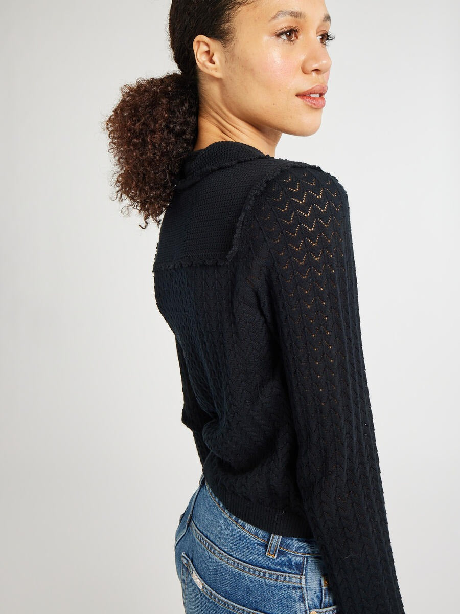 A person with curly hair is wearing a Mille Andie Cardigan Black with hand crochet trim and blue jeans. They are looking over their shoulder with a slight smile, against a plain light-colored background.