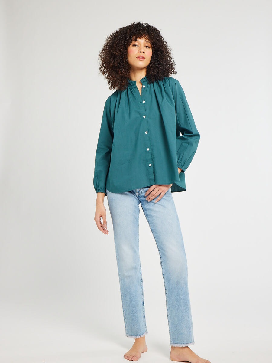 A person with curly hair stands against a plain background wearing the Mille Francesca Top Emerald Poplin from Mille and light blue jeans. They have one hand in their pocket and the other resting by their side.