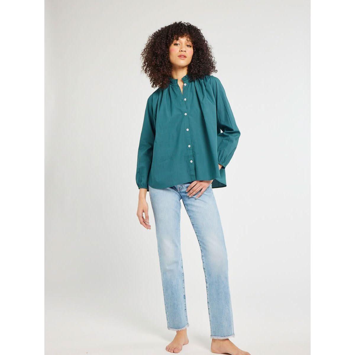 A person with curly hair stands against a plain background wearing the Mille Francesca Top Emerald Poplin from Mille and light blue jeans. They have one hand in their pocket and the other resting by their side.