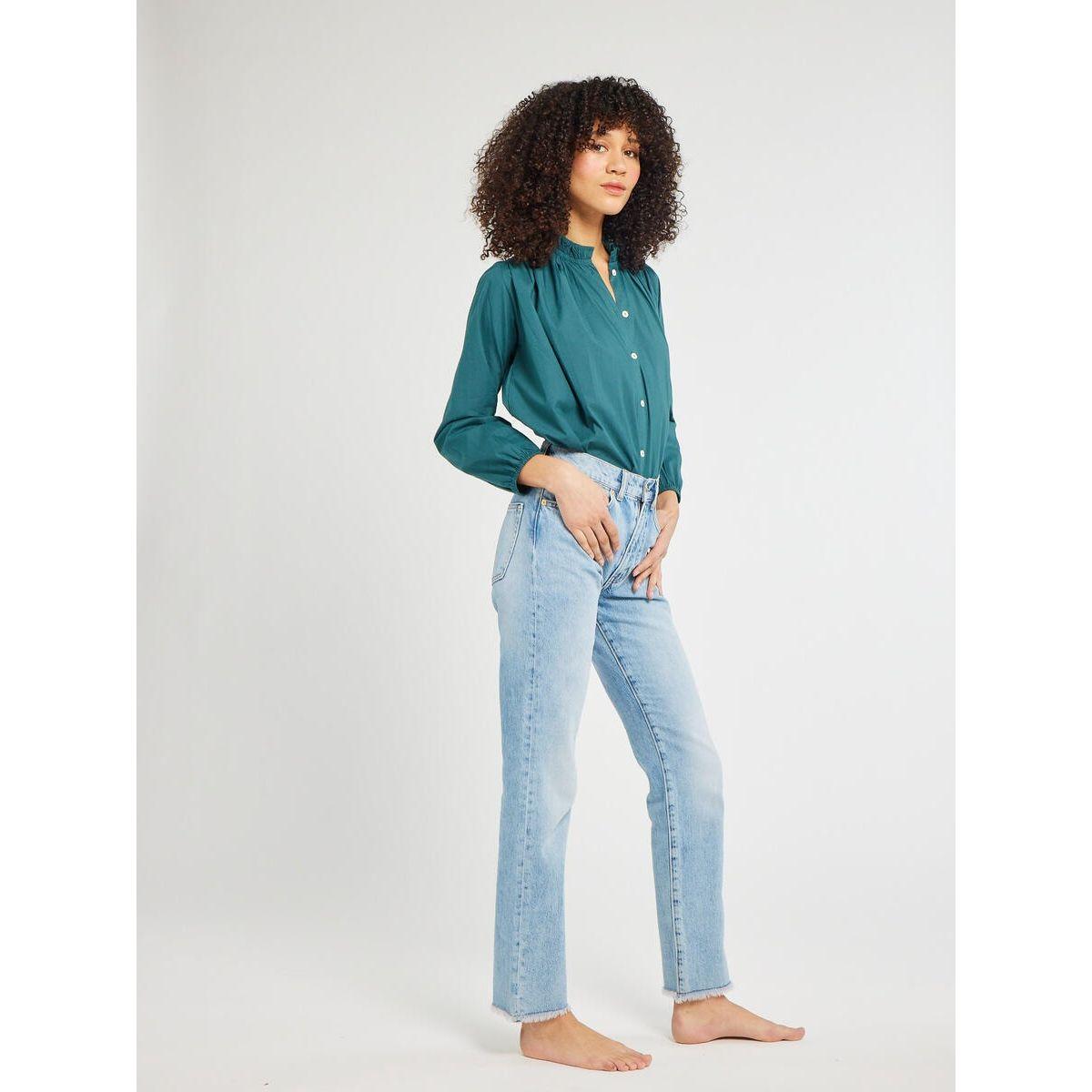 A person with curly hair stands barefoot against a plain background. They are wearing a relaxed silhouette, emerald poplin button-up shirt called the Mille Francesca Top from Mille and light blue high-waisted jeans. Their left hand is tucked into their pocket.