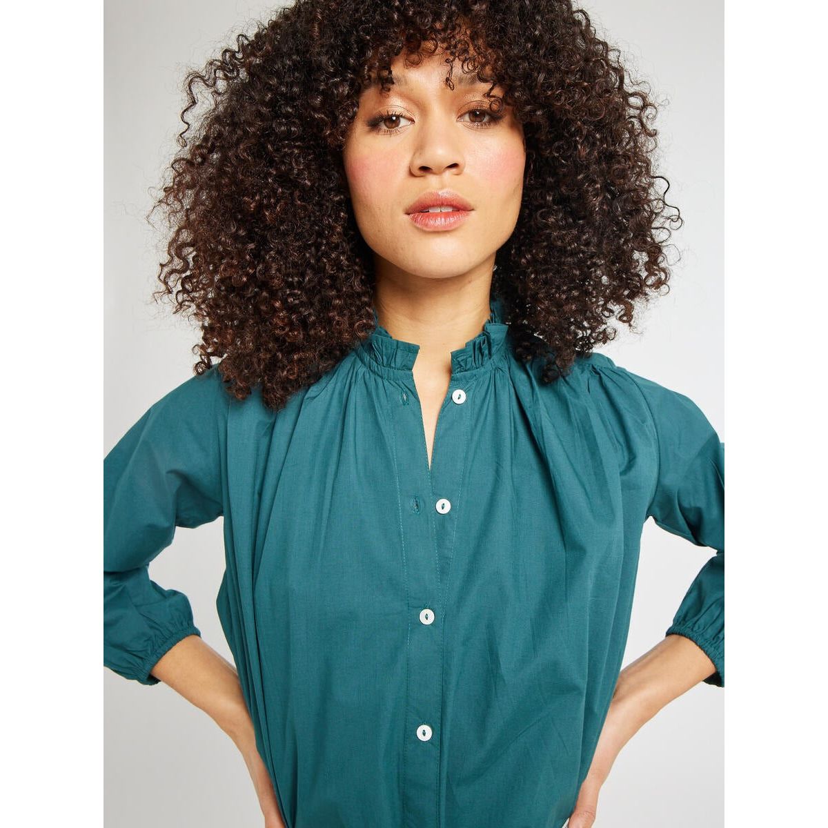 A person with curly hair is wearing the Mille Francesca Top Emerald Poplin by Mille. They are looking confidently at the camera with their hands on their hips. The background is neutral and light-colored.