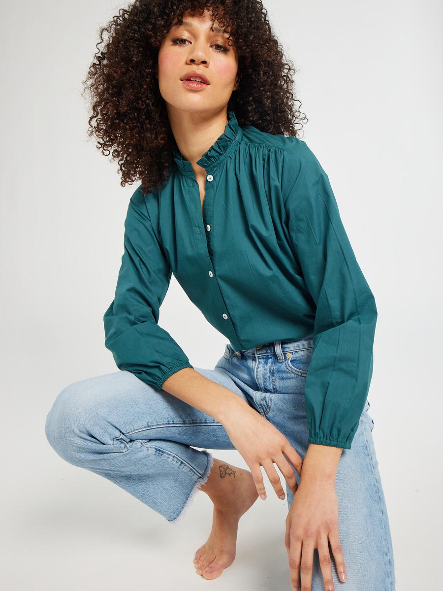 A person with curly hair is posing in the Mille Francesca Top Emerald Poplin by Mille and light blue jeans, kneeling on one knee in a neutral studio background.