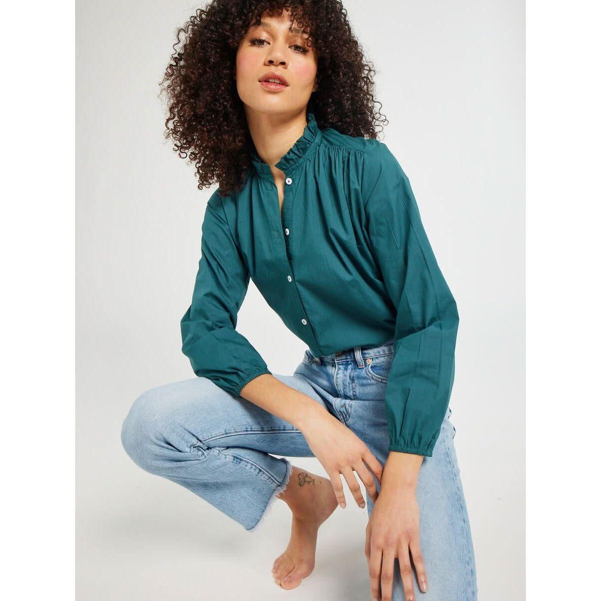 A person with curly hair is posing in the Mille Francesca Top Emerald Poplin by Mille and light blue jeans, kneeling on one knee in a neutral studio background.