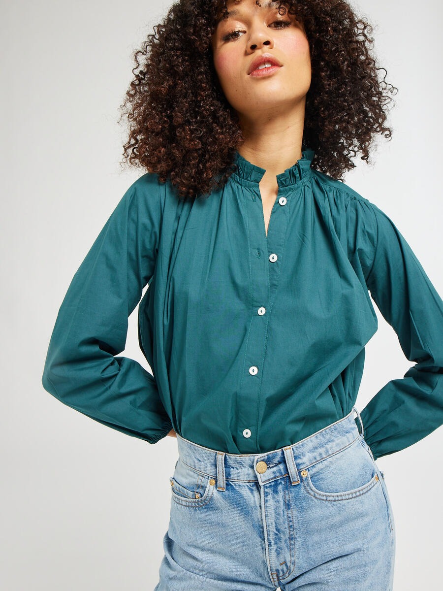 A person with curly hair poses confidently, wearing the Mille Francesca Top in Emerald Poplin paired with light blue high-waisted jeans. The background is a plain, light gray.