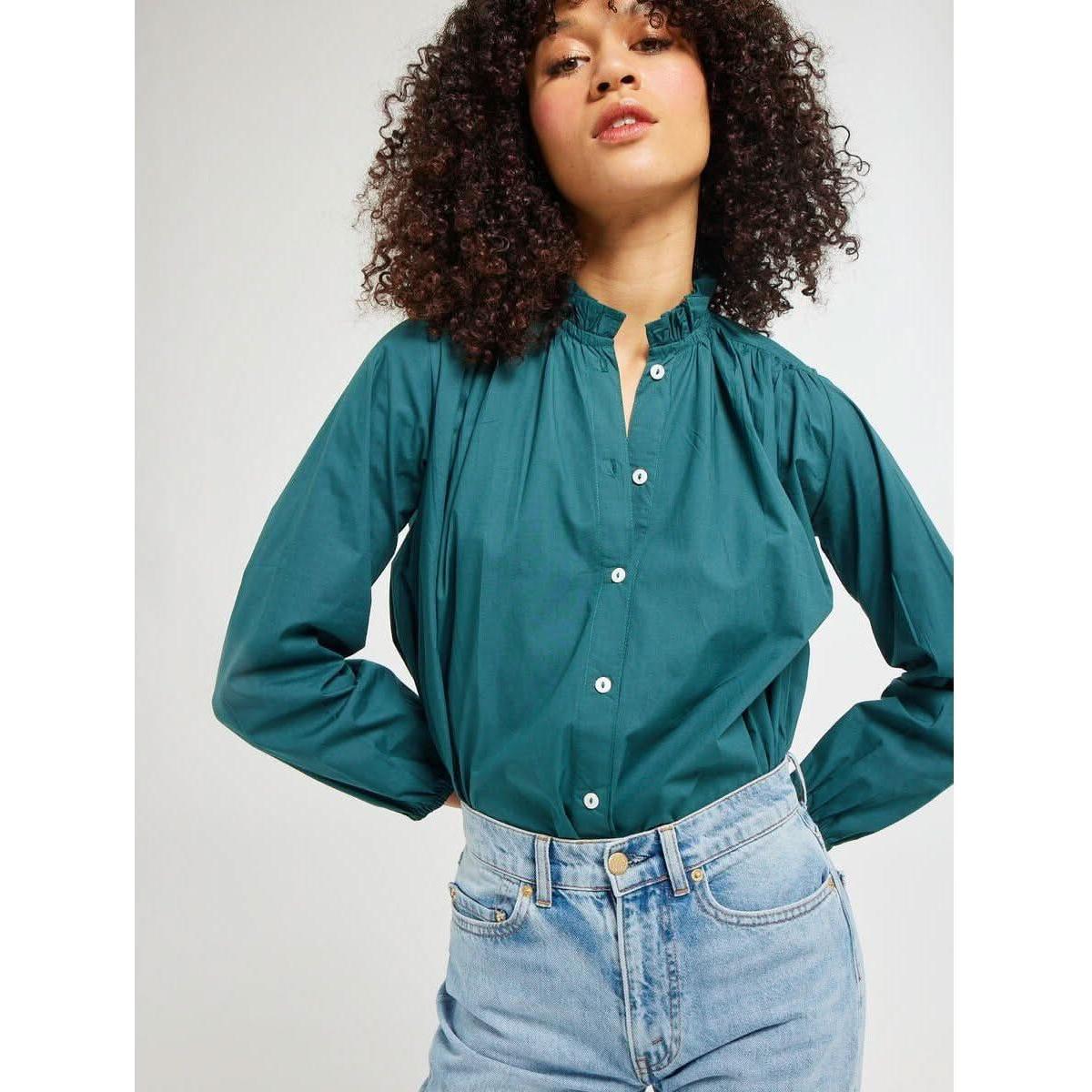 A person with curly hair poses confidently, wearing the Mille Francesca Top in Emerald Poplin paired with light blue high-waisted jeans. The background is a plain, light gray.