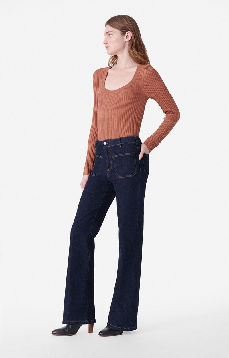 A person with long hair is modeling a rust-colored, long-sleeve, ribbed sweater and dark blue Vanessa Bruno Dompay Flare Jeans with a 70's flair. They are standing against a plain white background, looking to the side with one hand on their hip and the other hand in a pocket.