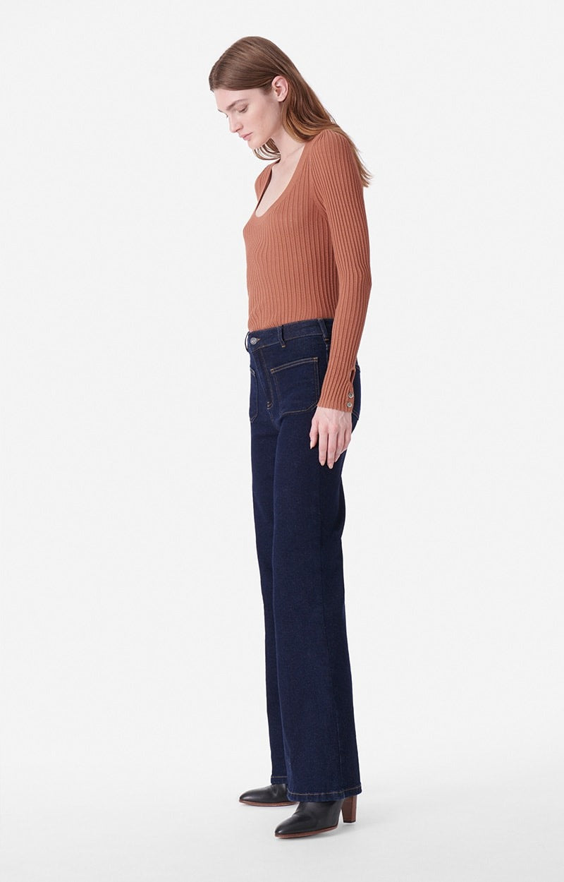 A woman stands in profile against a plain white background. She wears a ribbed, rust-colored long-sleeve top by Vanessa Bruno and high-waisted, dark blue Dompay flare jeans from Vanessa Bruno. She pairs the outfit with black ankle boots, her head slightly tilted downwards.