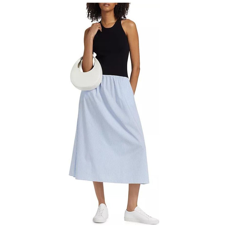 A person stands wearing the Naadam Mixed Media Midi Dress in a Black Blue Combo. They carry a white, crescent-shaped handbag and wear white sneakers. Their curly hair is slightly tousled, and one hand is on their hip. The background is plain white.