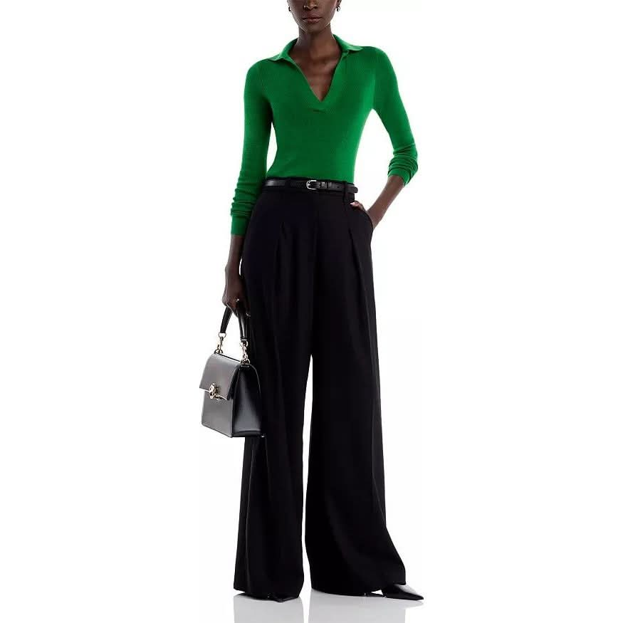 Wearing the Vanessa Bruno Dorita Top in green, made from soft cotton, and black high-waisted wide-leg trousers, a person complements their chic ensemble with a stylish black handbag. The look is elegantly enhanced by a black belt and pointed shoes as they pose against a white background.