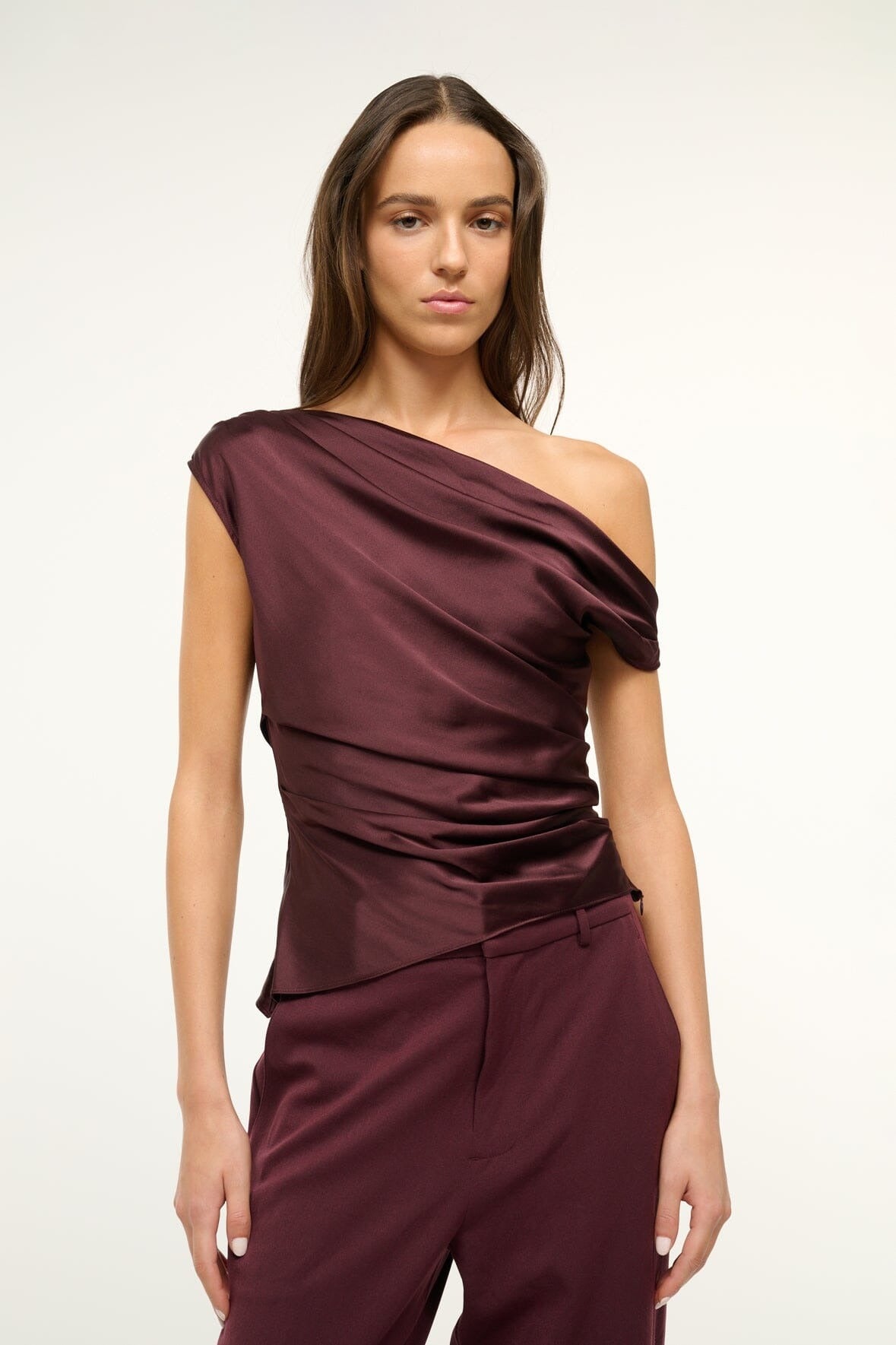 A person stands against a neutral background, wearing the Staud Phare Top in dark red. Complemented by matching pants, the silk satin fabric drapes elegantly. Their long, dark hair falls loosely around their shoulders as they maintain a calm expression, exuding effortless sophistication with this drapey fitted silhouette from Staud.