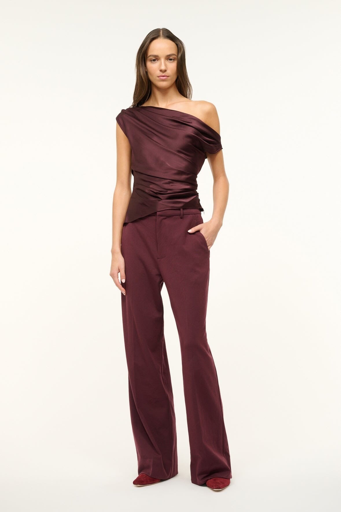 A model stands against a plain background wearing the stylish Staud Phare Top, an off-the-shoulder maroon silk satin blouse. It's paired with matching maroon wide-legged trousers. The outfit exudes elegance and sophistication, with the top's intricate draping and drapey fitted silhouette adding a touch of modern flair.