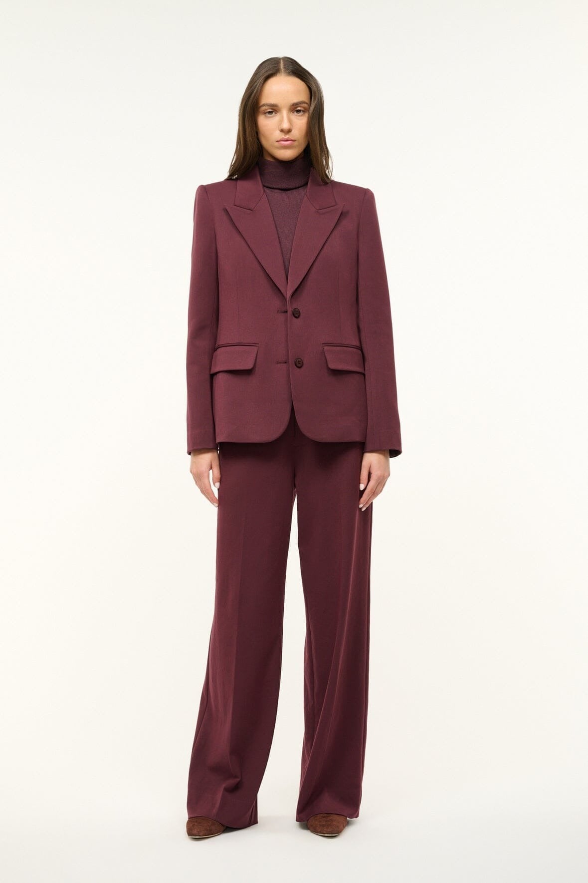 A person with long brown hair is standing against a plain light background. They are wearing the Staud Prince Pant Merlot—a burgundy suit paired with a matching turtleneck sweater and brown shoes. The flat front wide-leg pants feature back welt pockets, completing the tailored blazer ensemble by Staud. Their hands rest by their sides.