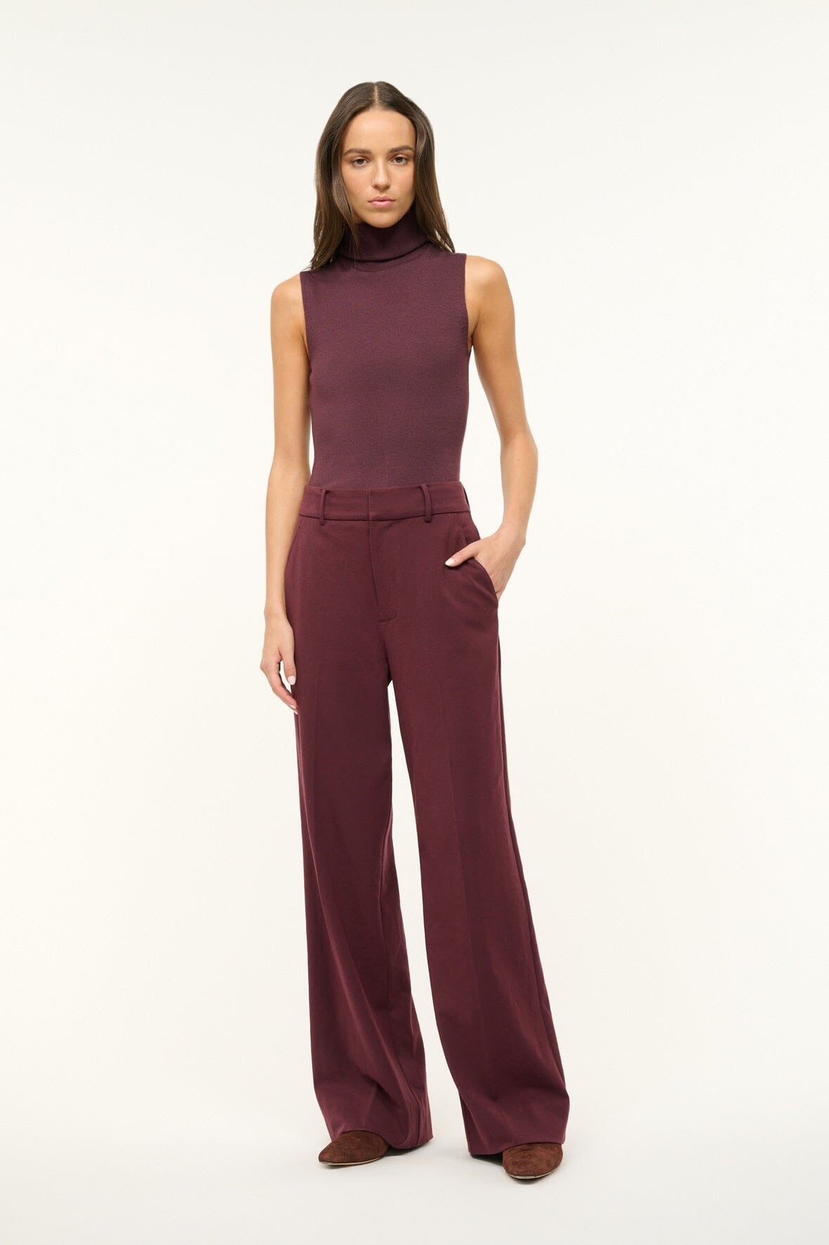 A woman stands against a plain background, wearing a sleeveless maroon turtleneck top and Staud's Prince Pant in Merlot, featuring back welt pockets and flat front wide legs. She has one hand in her pocket and is looking directly at the camera.