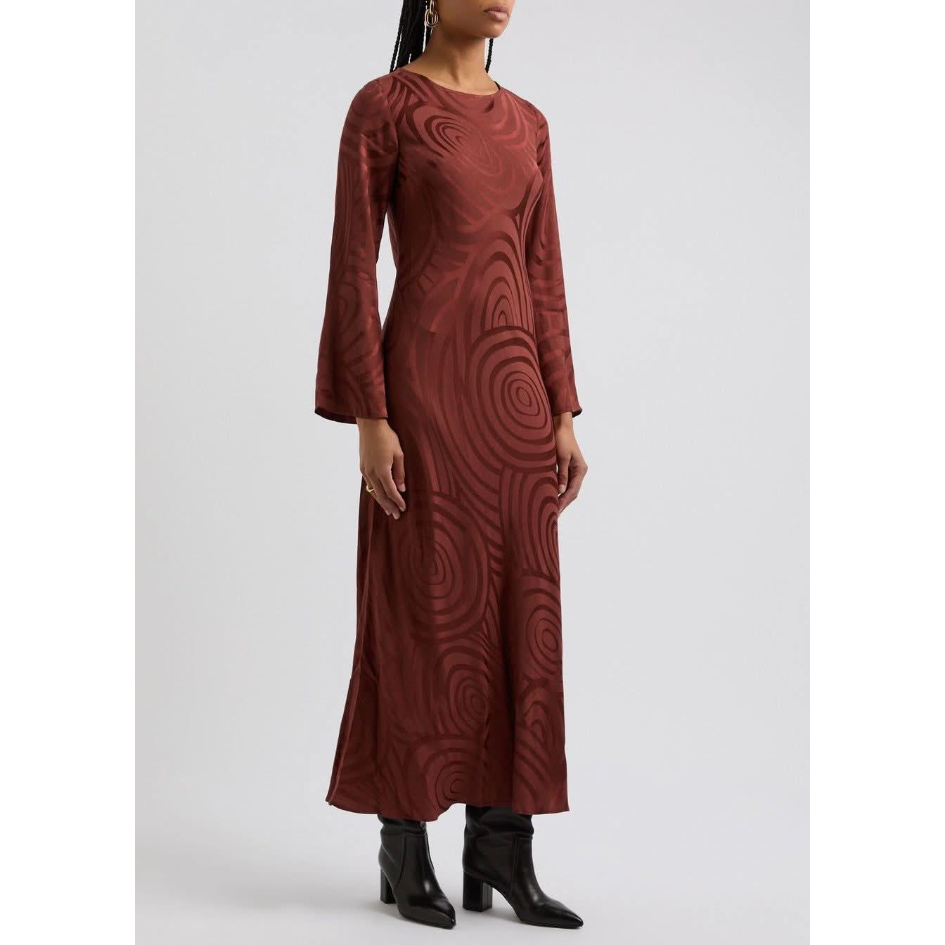 A person with braided hair is wearing a Rixo Alondra Dress Whirlpool Jacquard Merlot, featuring a brown, circular pattern on silk crepe de chine. They are accessorizing with earrings and a ring, and are standing in black heeled boots against a plain background.