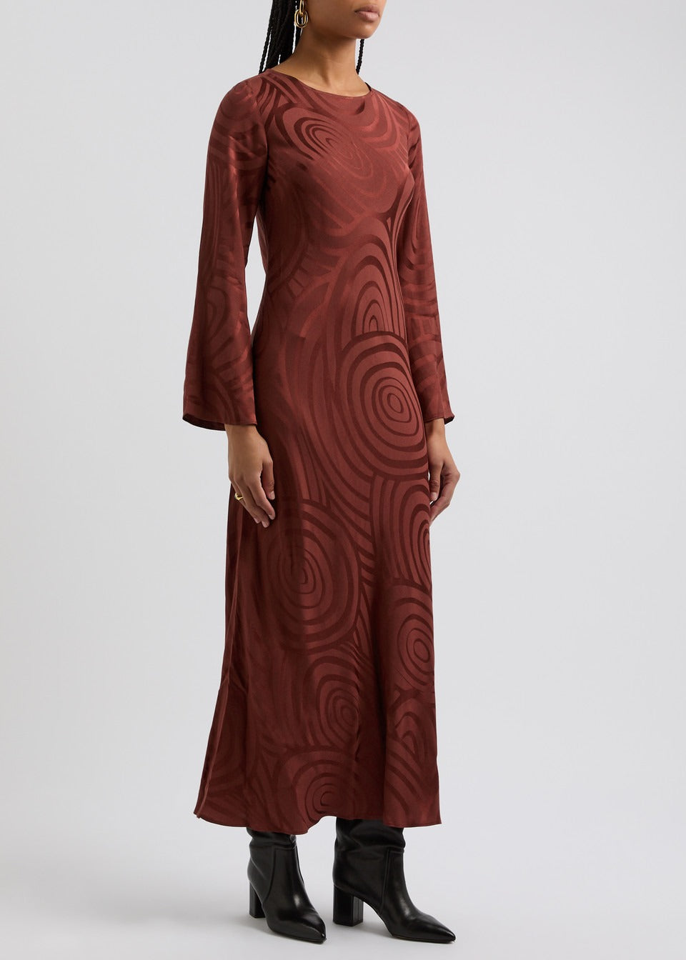 A person with braided hair is wearing a Rixo Alondra Dress Whirlpool Jacquard Merlot, featuring a brown, circular pattern on silk crepe de chine. They are accessorizing with earrings and a ring, and are standing in black heeled boots against a plain background.