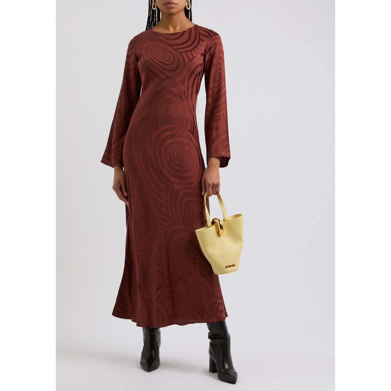 A person wearing a long-sleeved, ankle-length Rixo Alondra Dress in Whirlpool Jacquard Merlot is holding a small, light yellow handbag. The individual also wears black ankle boots. The background is plain and light-colored.