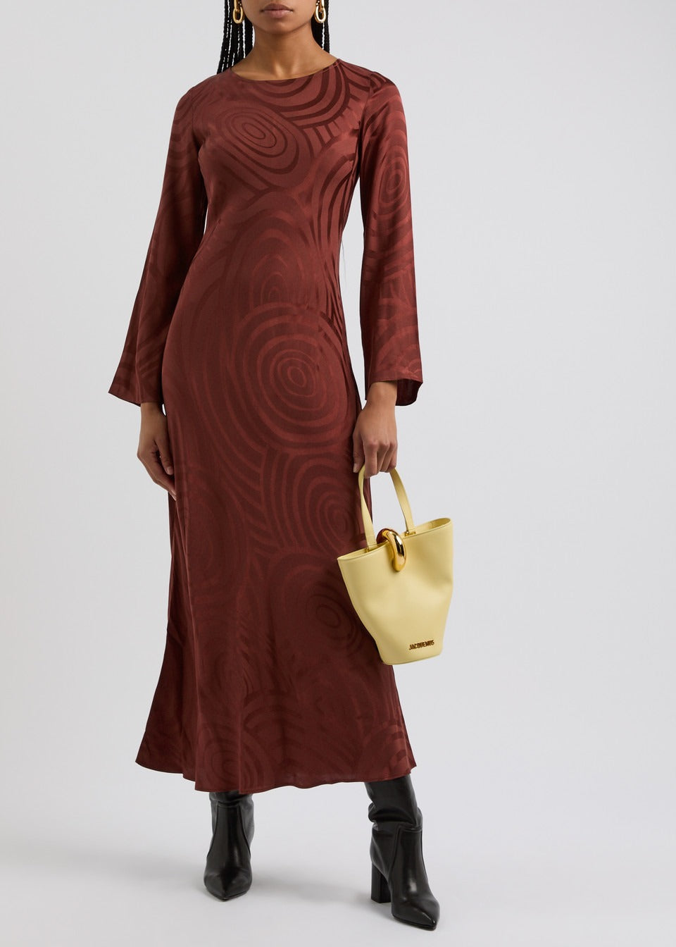 A person wearing a long-sleeved, ankle-length Rixo Alondra Dress in Whirlpool Jacquard Merlot is holding a small, light yellow handbag. The individual also wears black ankle boots. The background is plain and light-colored.