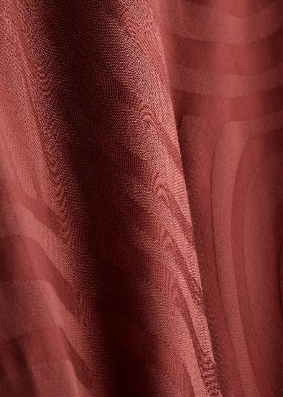 Close-up of the Whirlpool Jacquard Merlot fabric from the Rixo Alondra Dress. This textured material features a wavy pattern in a rich, reddish-brown color. The fabric has a subtle sheen, reminiscent of luxurious silk crepe de chine. The smoothly flowing lines create depth and movement, while the lighting enhances its undulating design.