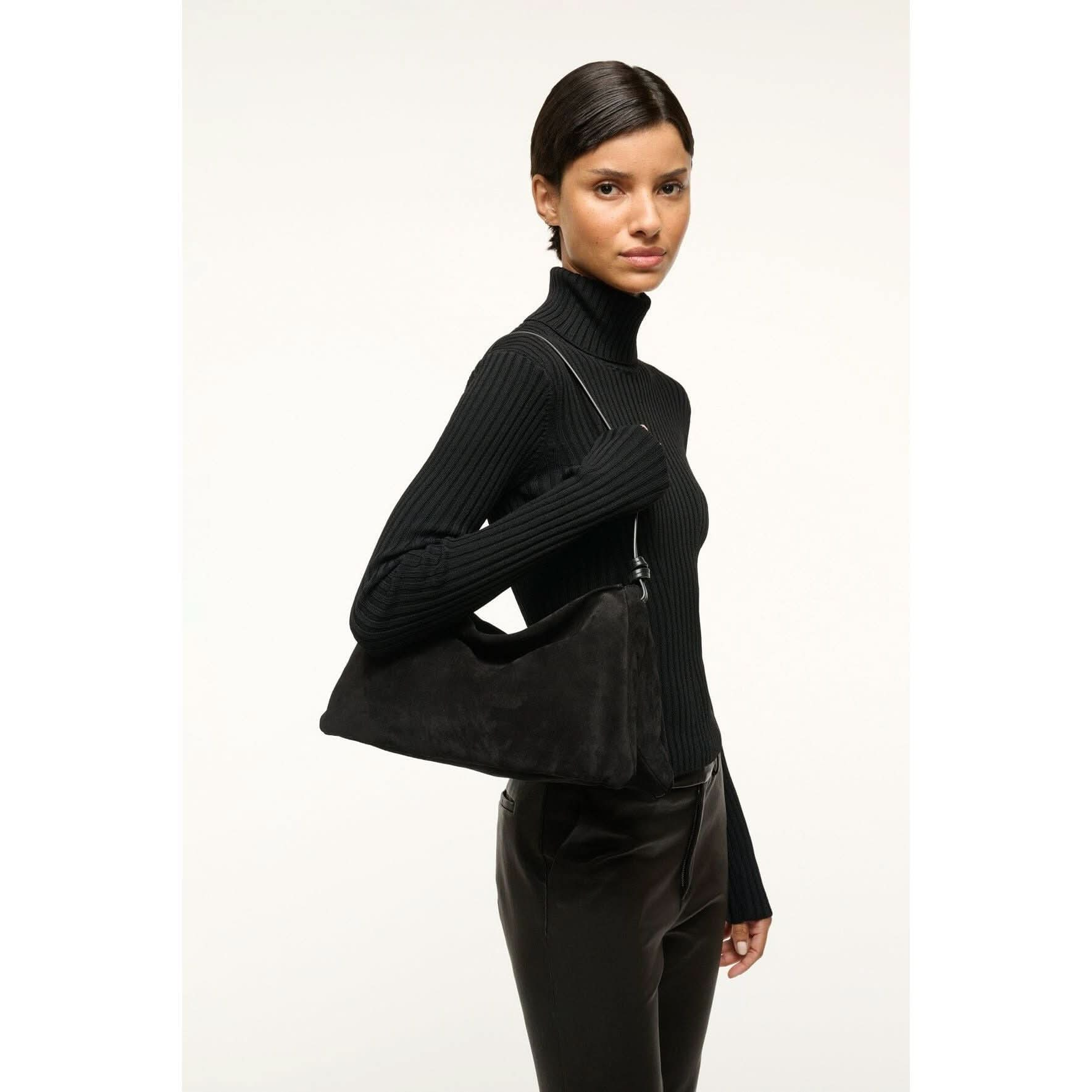 A person wearing a black turtleneck sweater and leather pants holds the chic Staud Wally Bag in black pebbled leather over their shoulder against a plain background.