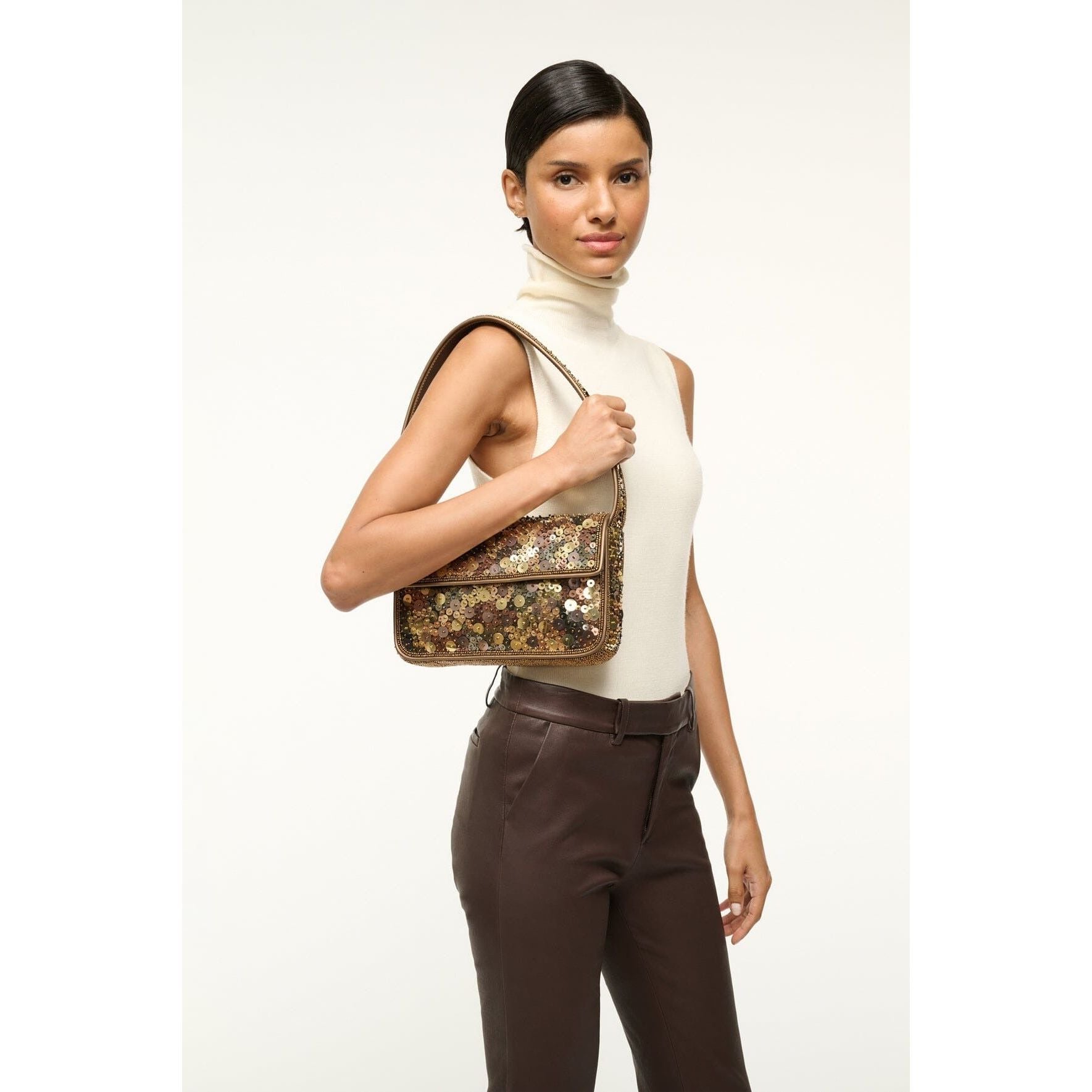 A person with short dark hair is wearing a sleeveless turtleneck top and brown pants, holding the Staud Tommy Bag in Gilded Sequins with a sequined floral design. The background is plain white.