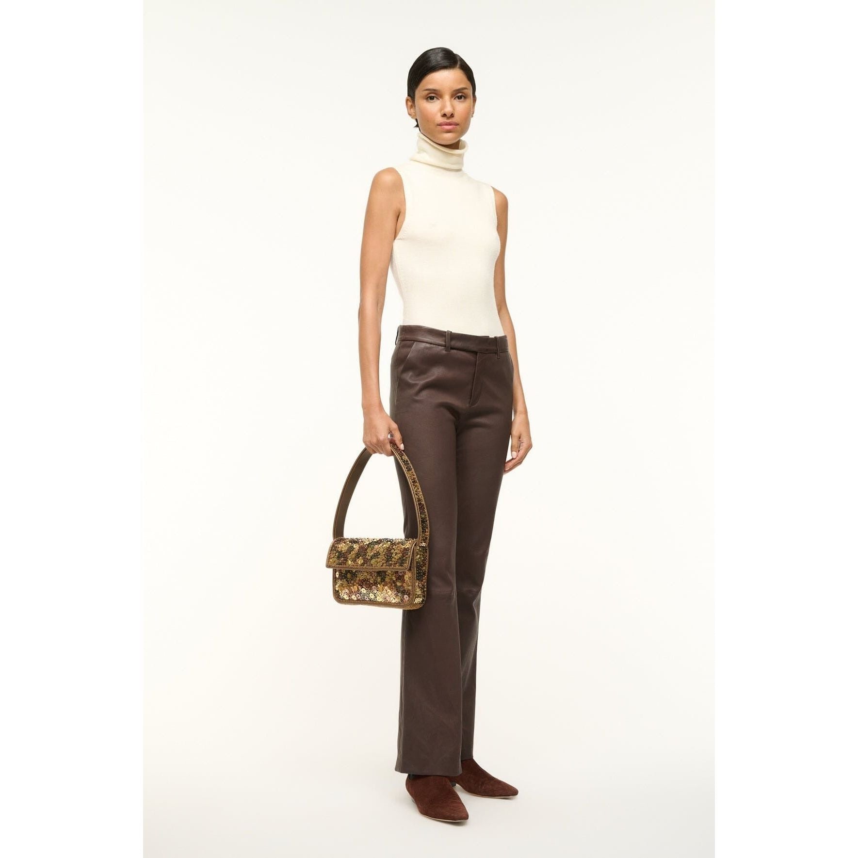 A person stands against a plain background wearing a sleeveless white turtleneck and brown trousers, holding the Staud Tommy Bag in Gilded Sequins with a floral beaded design. They have short hair and are wearing vegan leather brown shoes.