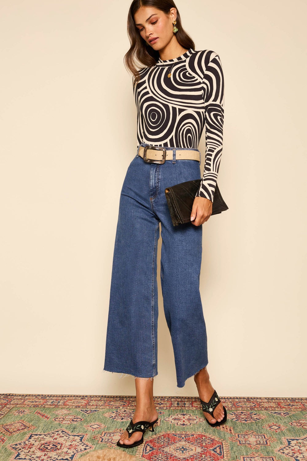 A woman with wavy hair wears the Rixo Tully Top Whirlpool Mono, a black and white geometric patterned long-sleeve blouse from Rixo. She pairs it with high-waisted wide-leg blue jeans and black heeled sandals. The fitted silhouette of her outfit is accentuated by the stretchy jersey material of the top. She holds a black clutch and stands on a colorful patterned rug against a plain background.