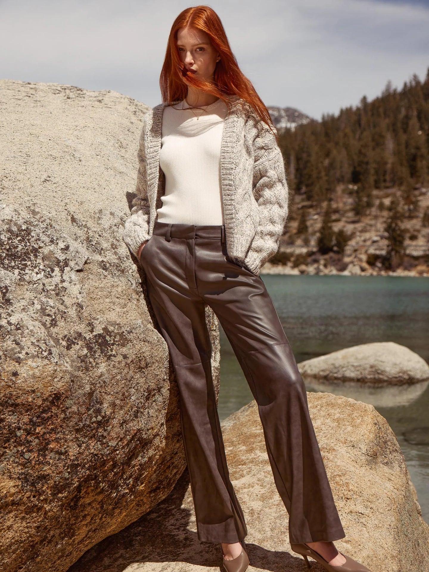 A person with long red hair stands outdoors against large rocks and a scenic backdrop of a lake and forested mountains. They are wearing a white top, a Brochu Walker knitted cardigan, and dark Brochu Walker Namari Vegan Leather Pants, with their hands in their pockets and looking directly at the camera.