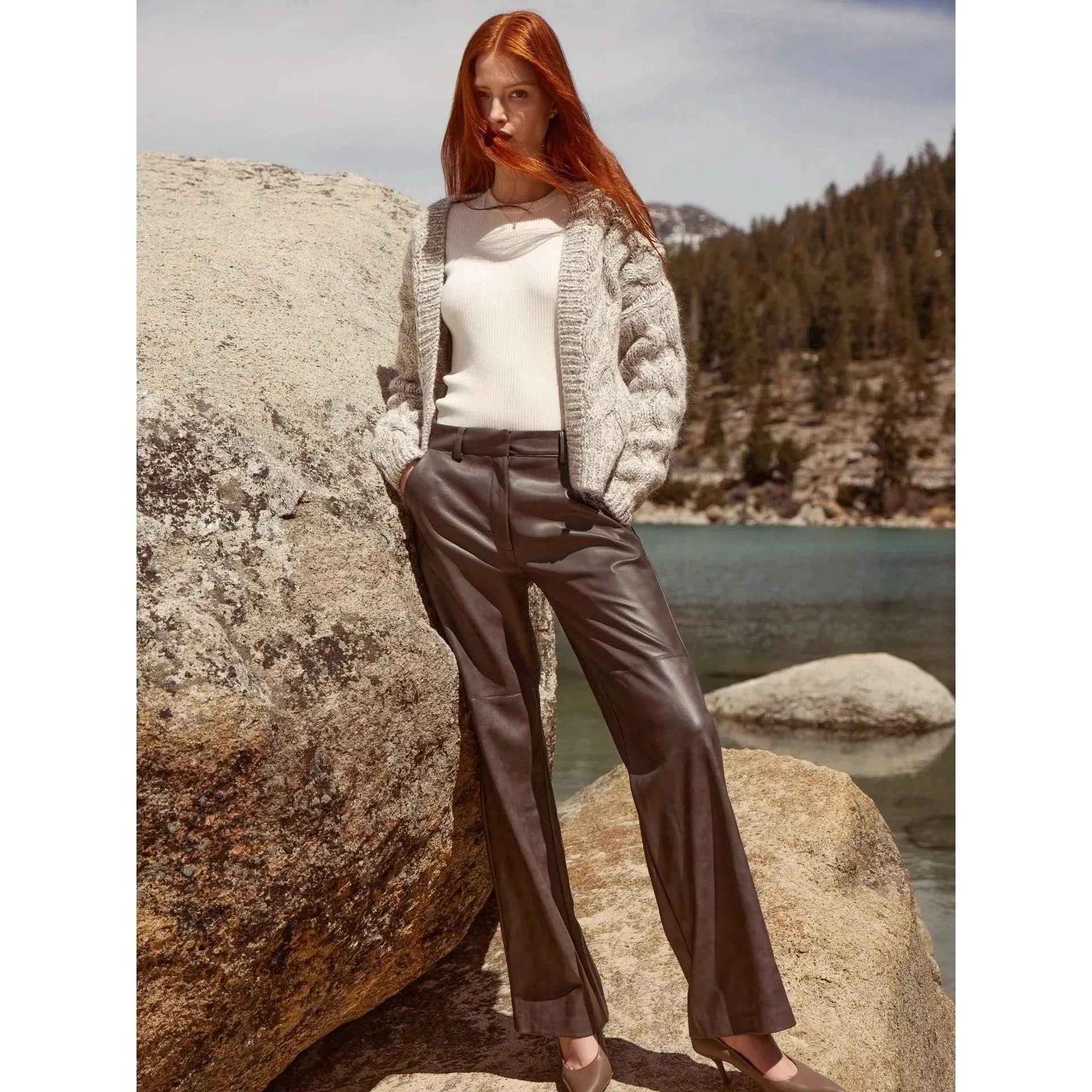 A person with long red hair stands outdoors against large rocks and a scenic backdrop of a lake and forested mountains. They are wearing a white top, a Brochu Walker knitted cardigan, and dark Brochu Walker Namari Vegan Leather Pants, with their hands in their pockets and looking directly at the camera.