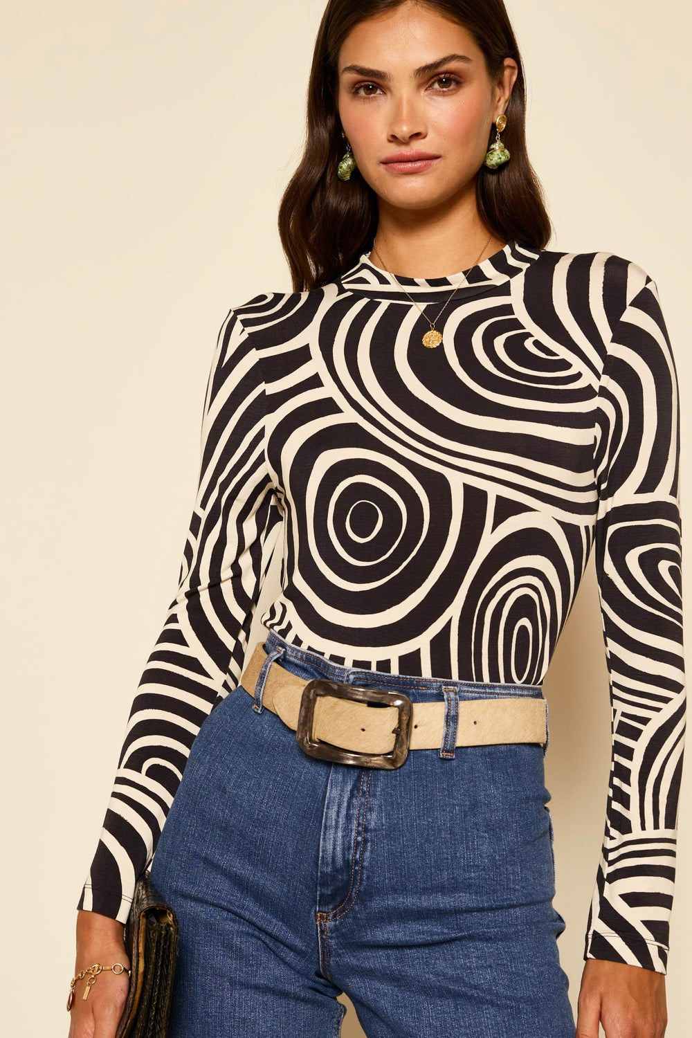 A woman with long dark hair is wearing the Rixo Tully Top Whirlpool Mono, a long-sleeve, black-and-white, abstract patterned top made from stretchy jersey. She has blue jeans with a beige belt, green earrings, and a gold necklace. She is holding a clutch and standing against a beige background.