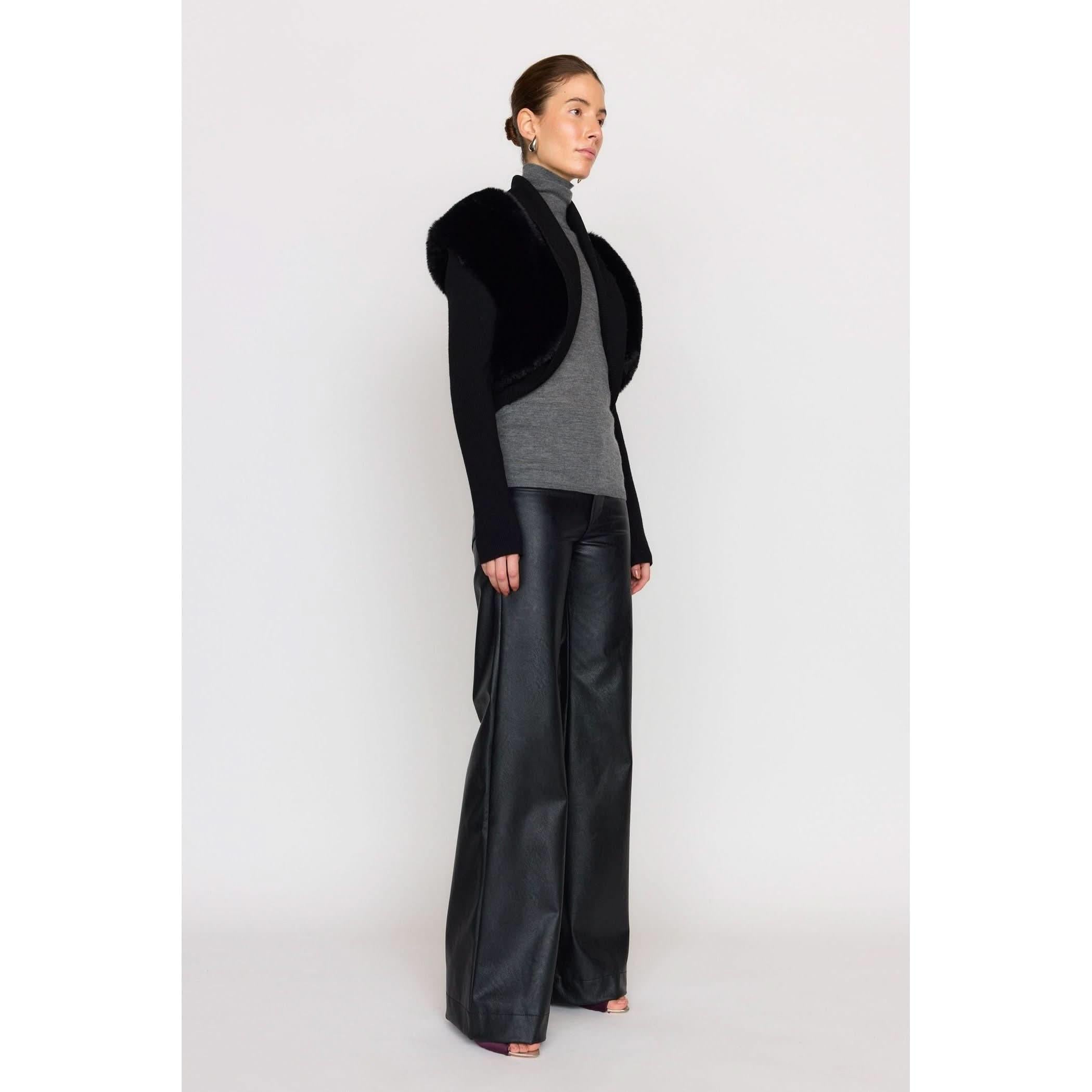 A person stands against a plain background wearing the Christy Lynn Criselle Knit in black, complemented by a black fur shrug and wide-legged vegan leather pants. They exude elegance and confidence with their hair styled in a neat bun.