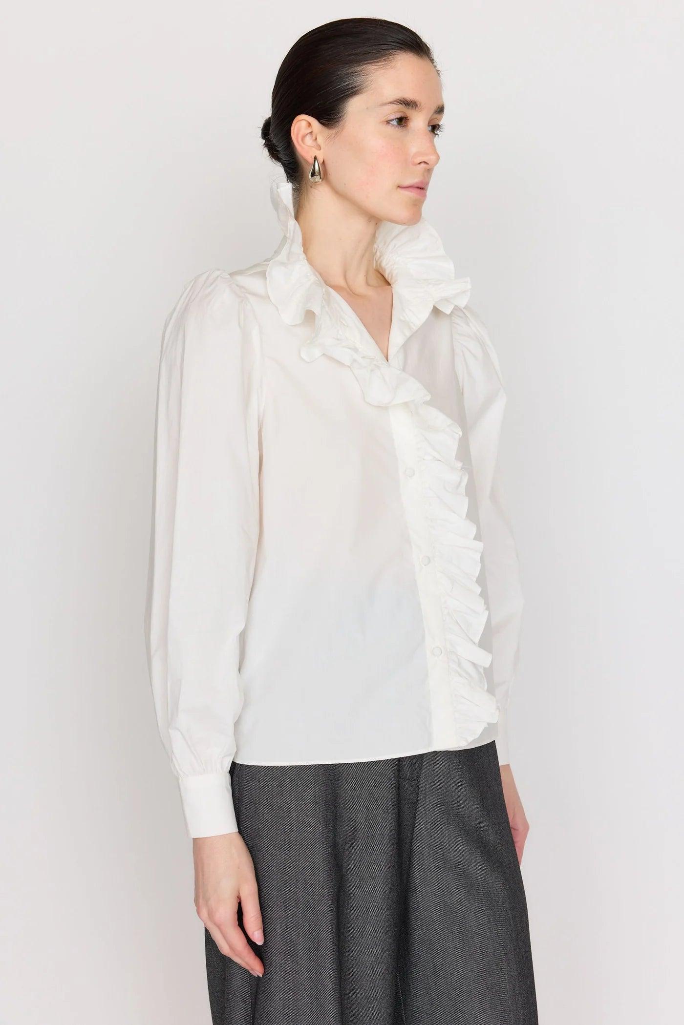 A woman with dark hair tied back is posing against a plain background. She is wearing the Christy Lynn Medora Top, a long-sleeve white blouse from Christy Lynn made of cotton poplin, featuring ruffled details along the collar and down the front, paired with high-waisted gray trousers. Small hoop earrings adorn her ears, and she appears contemplative.