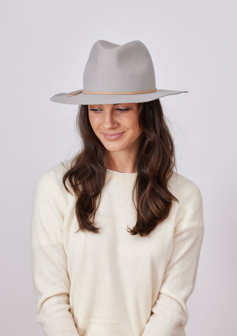 A person with long, wavy brown hair is wearing a Hat Attack Classic Amelia in Light Grey, featuring a tan band. They are dressed in a cream-colored sweater and are looking downward with a slight smile. The plain, light-colored background highlights their cozy style, ideal for pairing with cotton denim on casual days from the Hat Attack collection.