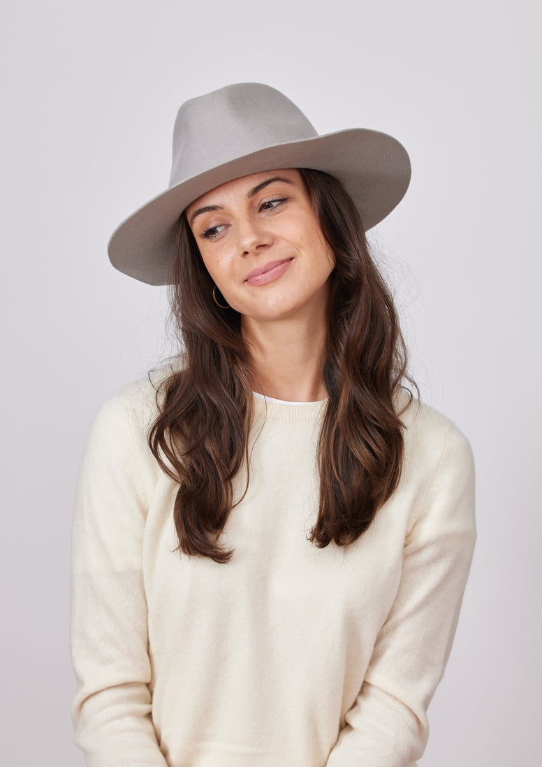 A person with long, wavy brown hair is wearing the Hat Attack Classic Amelia in light grey — a wide-brimmed hat that offers SPF protection. They are also dressed in a cream-colored sweater. With a gentle smile on their face, they gaze slightly to the side against a plain, light grey background.