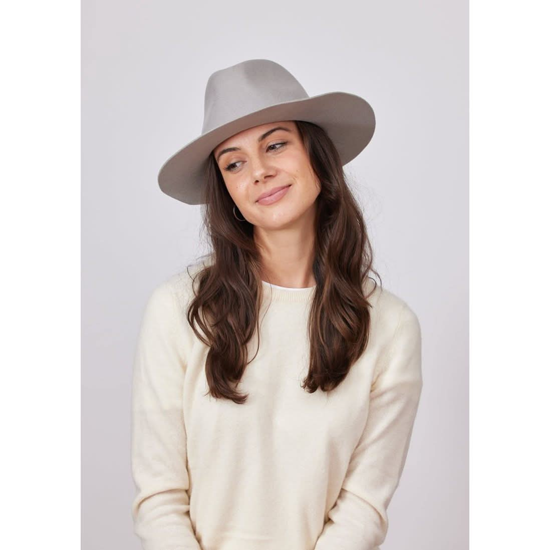 A person with long brown hair is wearing the Hat Attack Classic Amelia in light grey and a cream sweater, looking down with a slight smile against a plain light gray background.