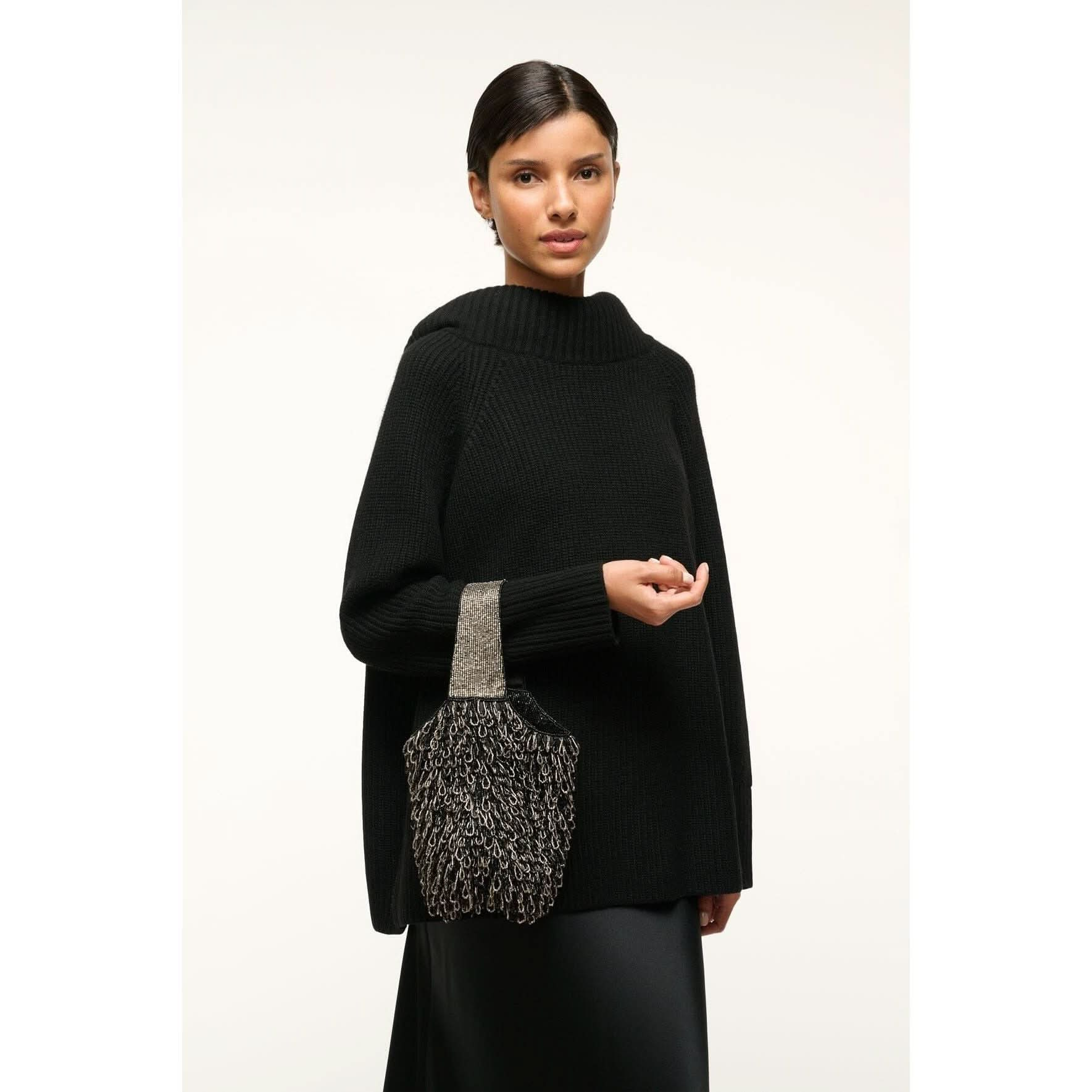 A person wearing a black sweater and skirt holds the Staud Haruko Beaded Bag, featuring a chain pattern and silver handle, against a plain background.