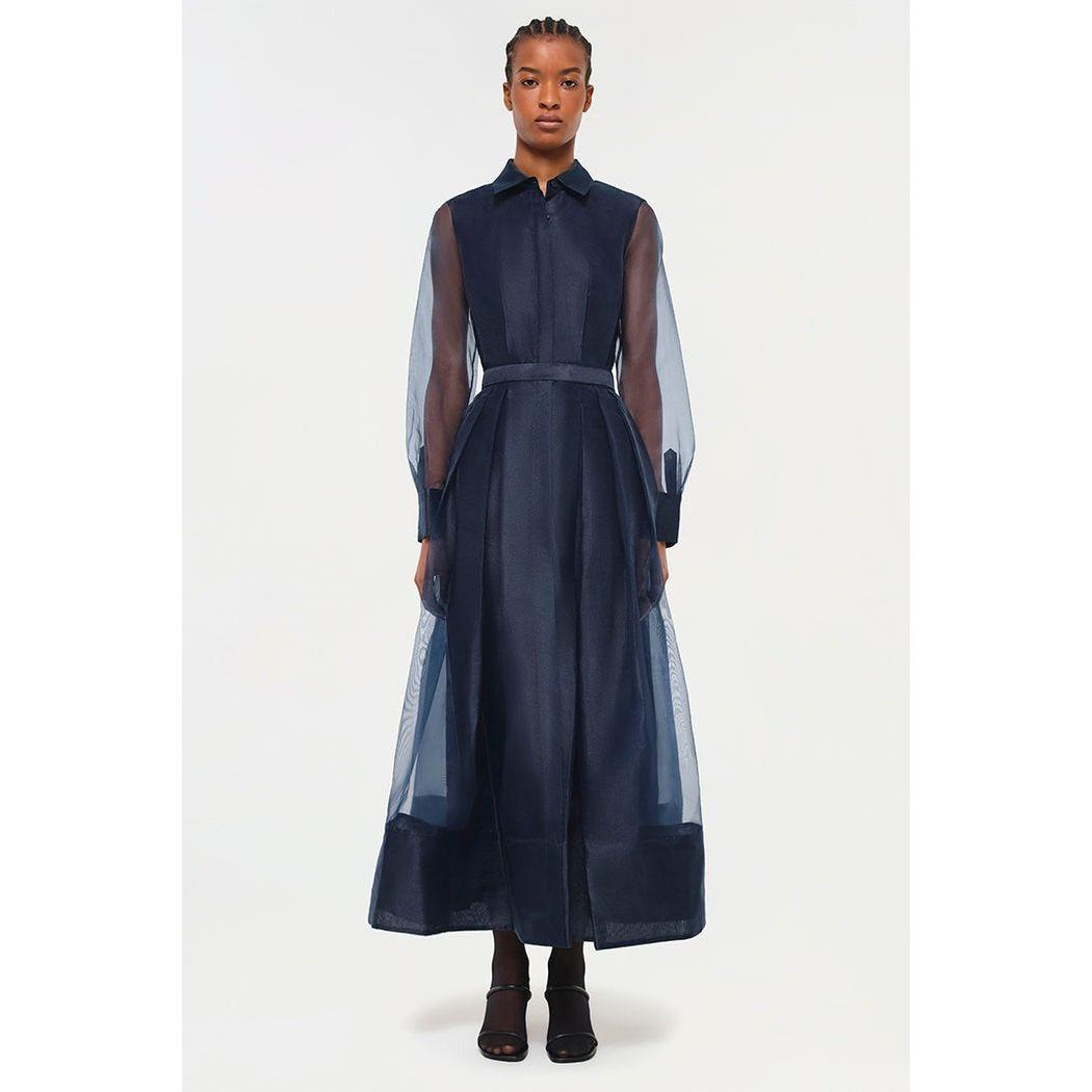 A person stands elegantly in the Simkhai Montgomery Button Up Dress, a long, navy blue ensemble featuring sheer sleeves and a collar. The fitted waist and pleated skirt add sophistication against the plain white background.