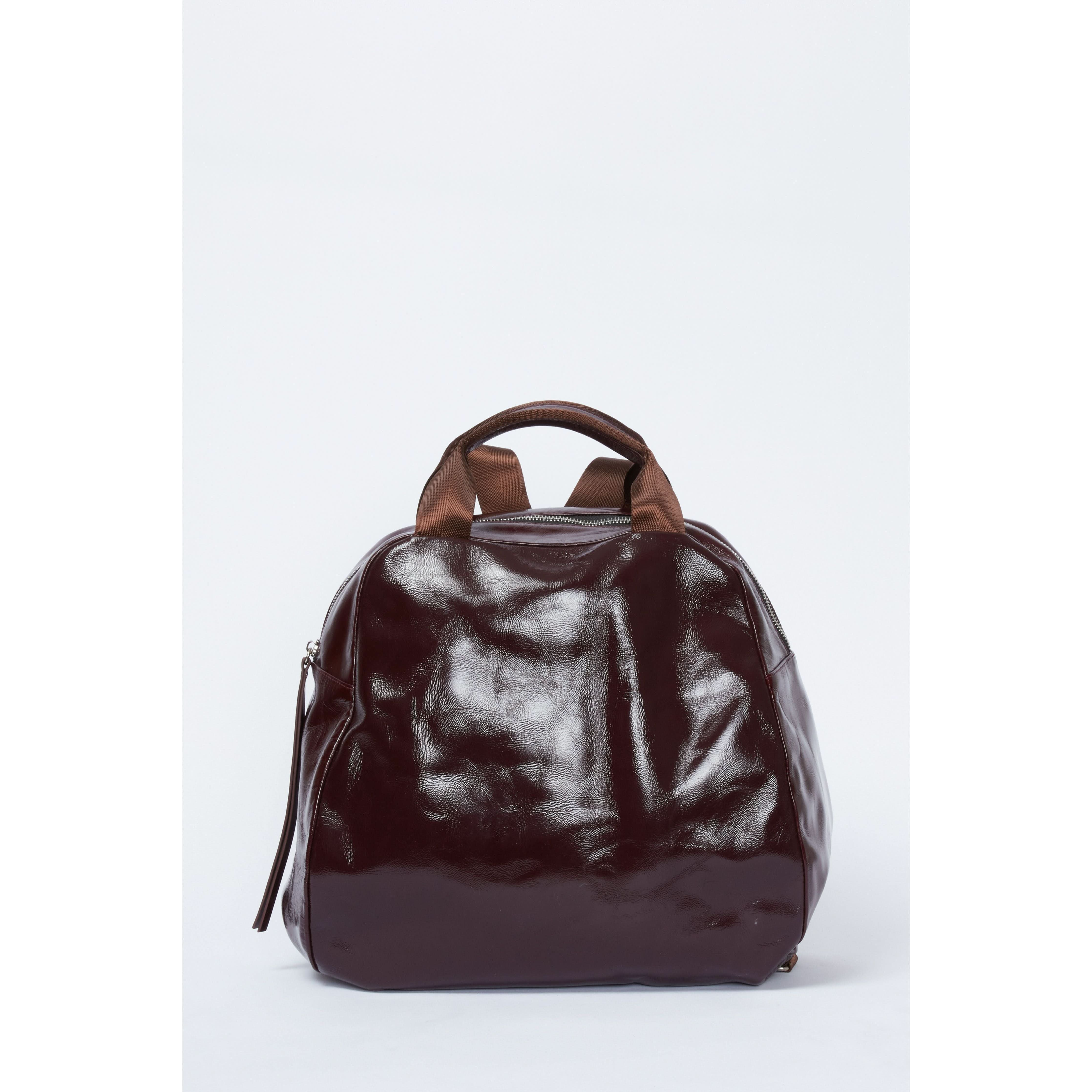 The Rachel Comey Lynbrook Backpack in Cherry is a medium-sized, glossy dark brown crinkled patent leather bag with a rounded shape and two short top handles. It features an eye-catching silver zipper and a small side pocket, alongside adjustable shoulder straps for convenience, set against a plain white background.