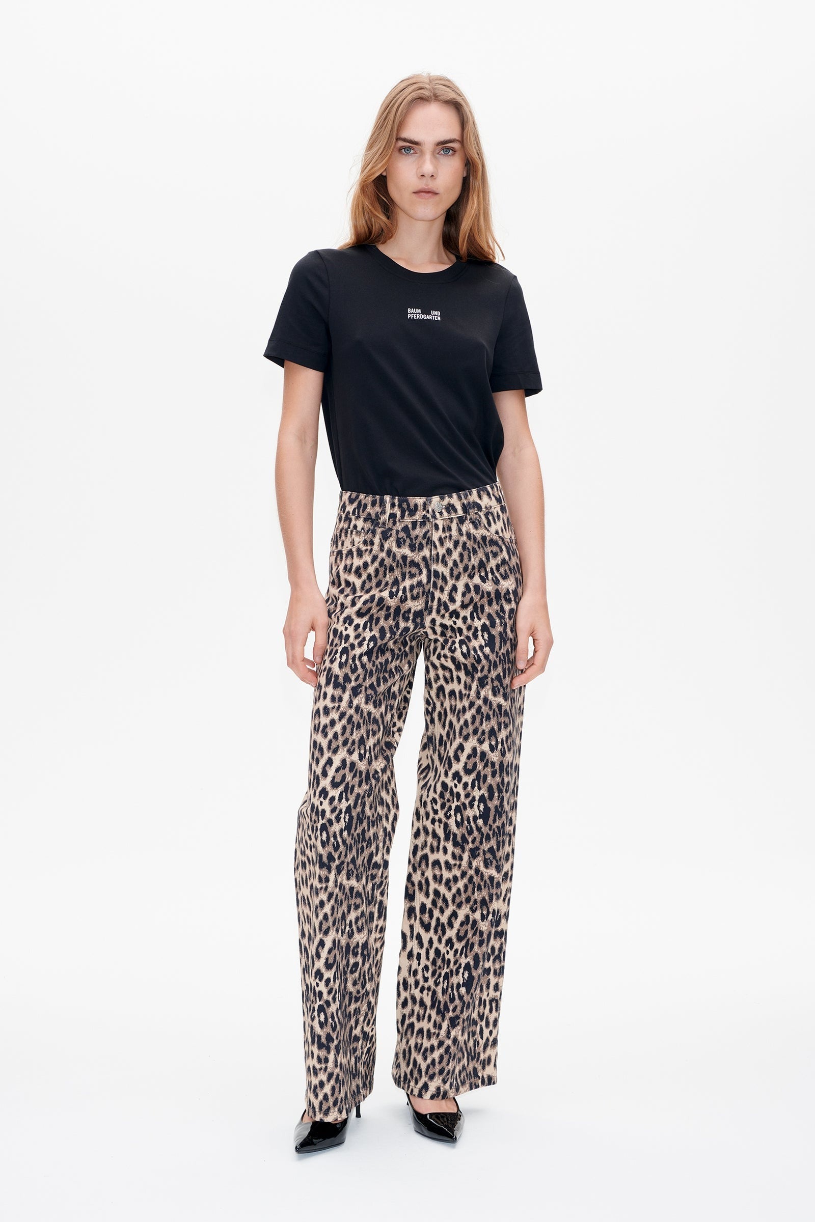 A woman stands against a plain white background, showcasing the Baum und Pferdgarten Nicette Jean by Baum und Pferdgarten. She is styled in a black t-shirt along with high-waisted, loose-fitting leopard print pants made from organic cotton, paired with black high-heeled shoes. She has long hair and a neutral expression.