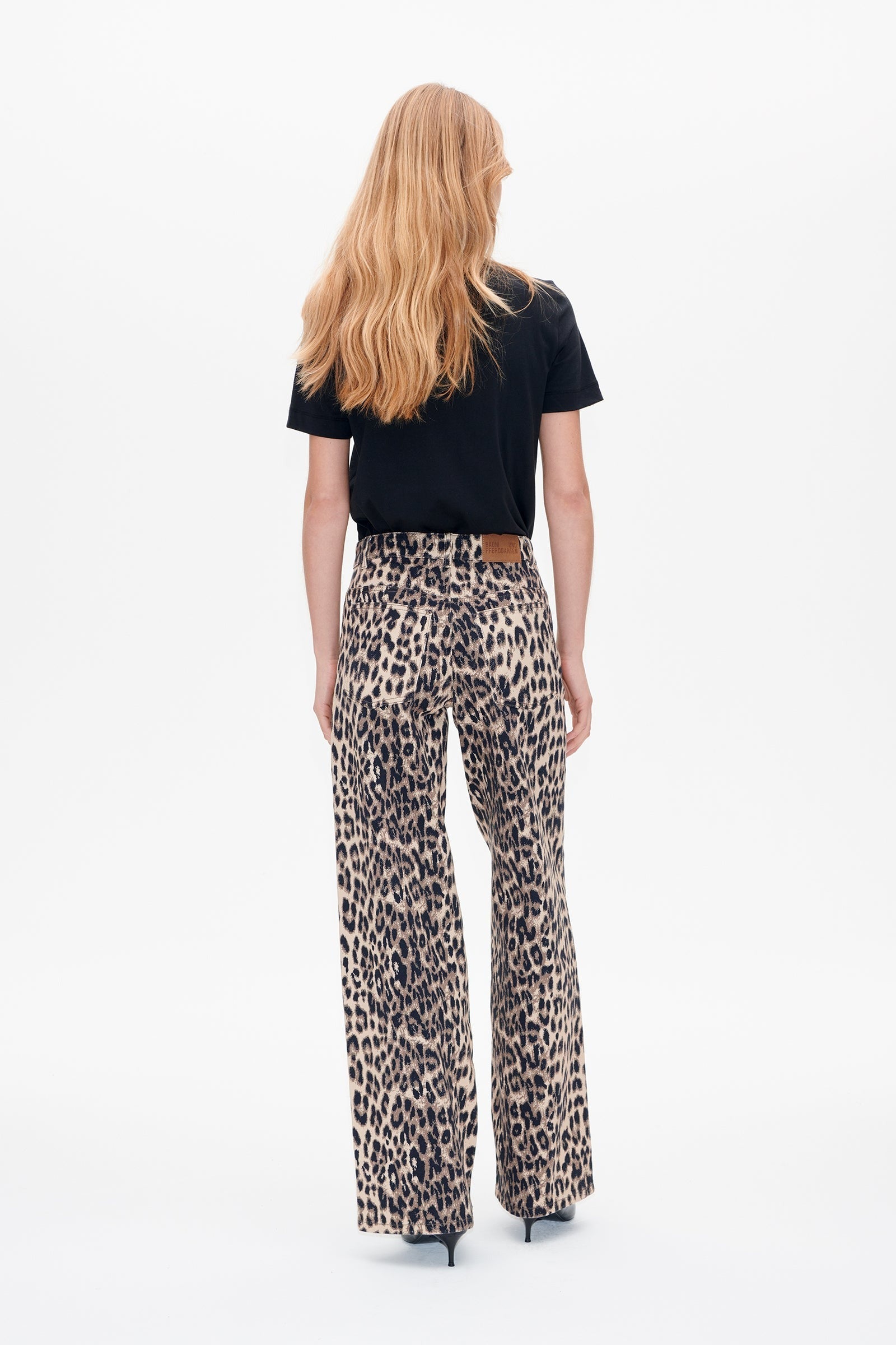 A person with long blond hair is standing with their back facing the camera. They are wearing a black T-shirt and Baum und Pferdgarten Nicette Jeans, which feature a leopard print wide-leg design made from organic cotton. They are also wearing black high-heeled shoes. The background is plain white.