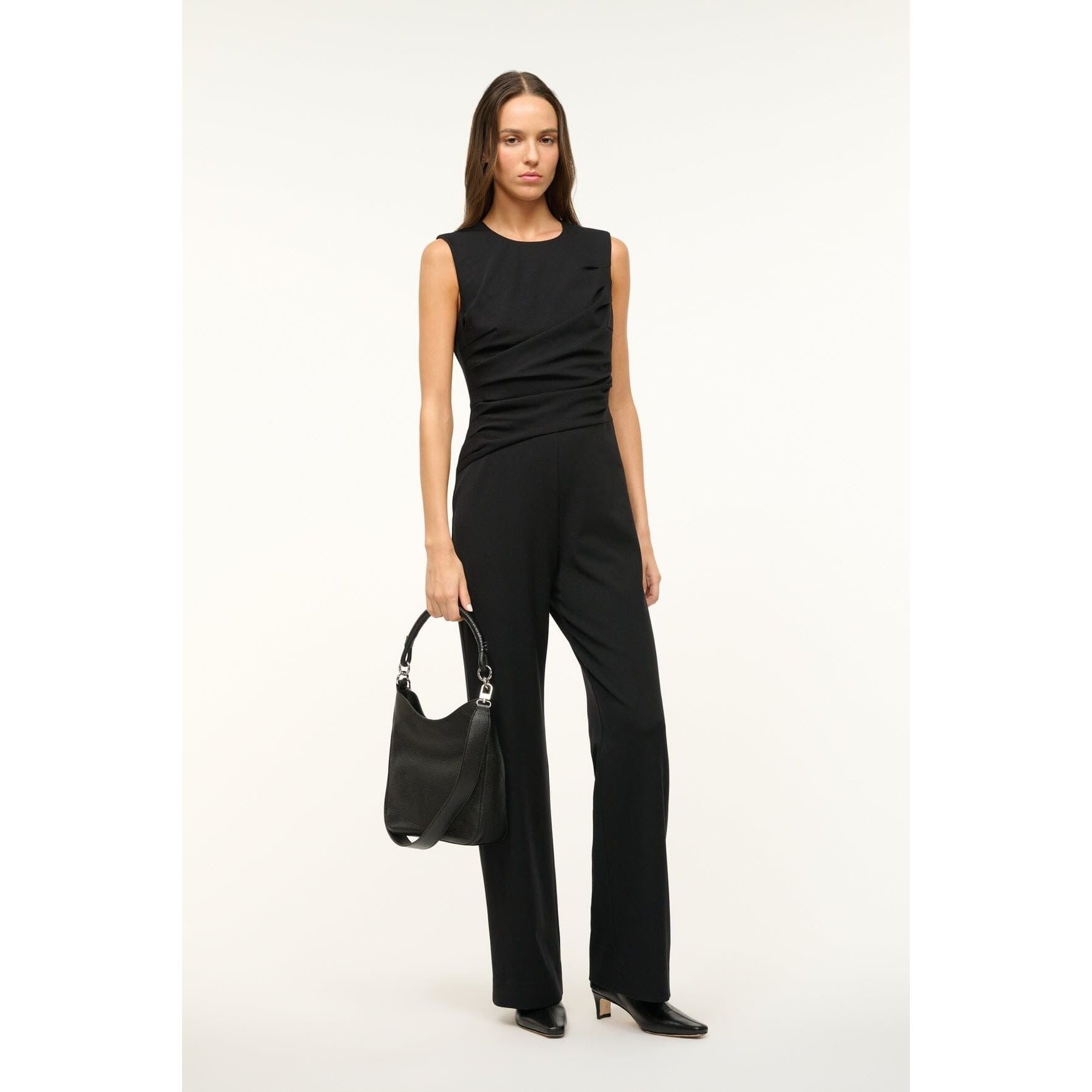 A chic woman in a sleeveless black jumpsuit holds the Staud Mel Bag Black, crafted from sleek cow leather and featuring a removable crossbody strap. Her long dark hair cascades elegantly as she steps confidently in black heeled shoes. The minimalist light gray background accentuates her timeless style.