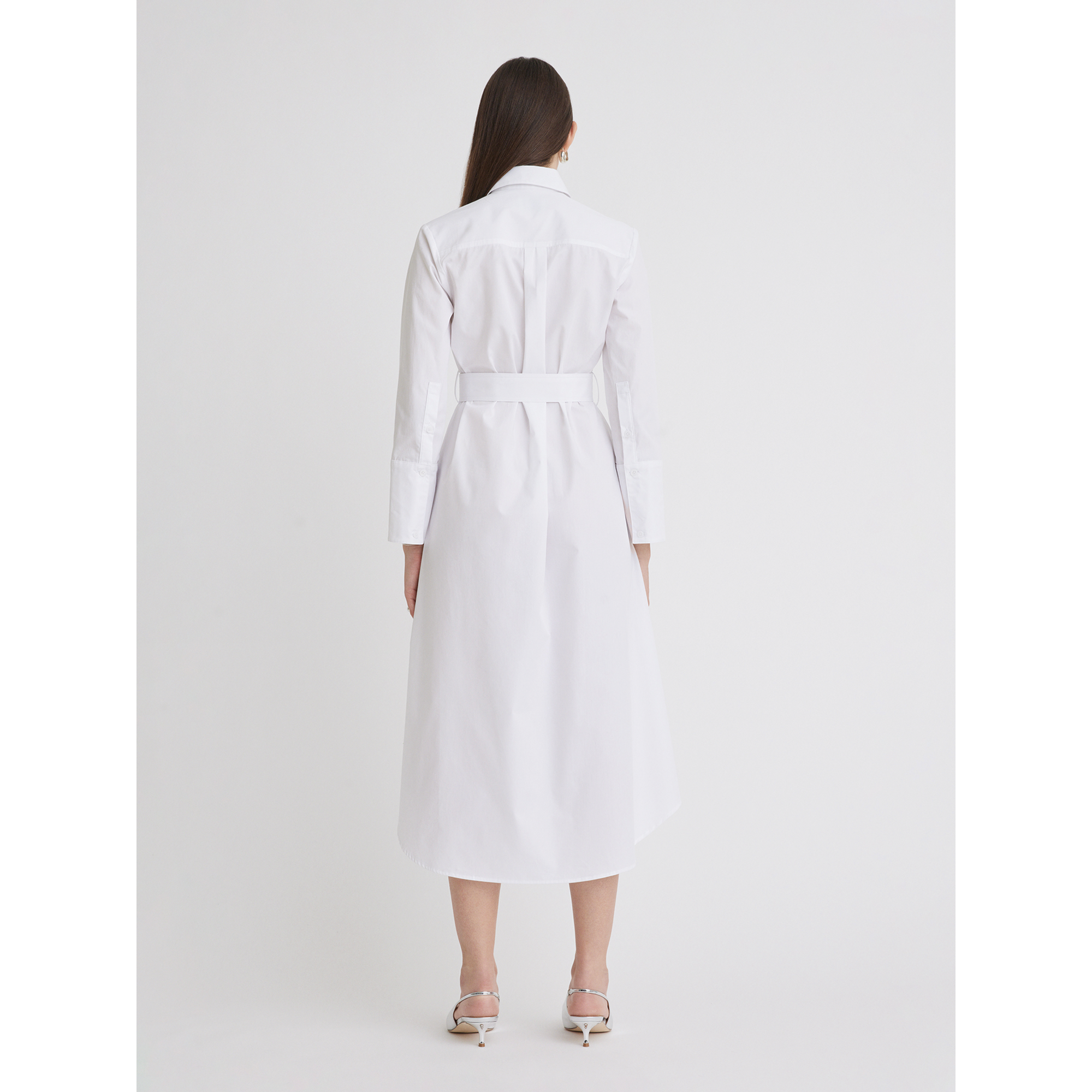 A woman with straight brown hair is standing and facing away from the camera. She is wearing a Matthew Bruch Cargo Button Up Midi White Poplin Shirt Dress, a 100% cotton, long-sleeved, belted design crafted in the USA. Complementing her outfit are white heeled sandals, all set against a plain white background. This elegant piece should be dry cleaned only.