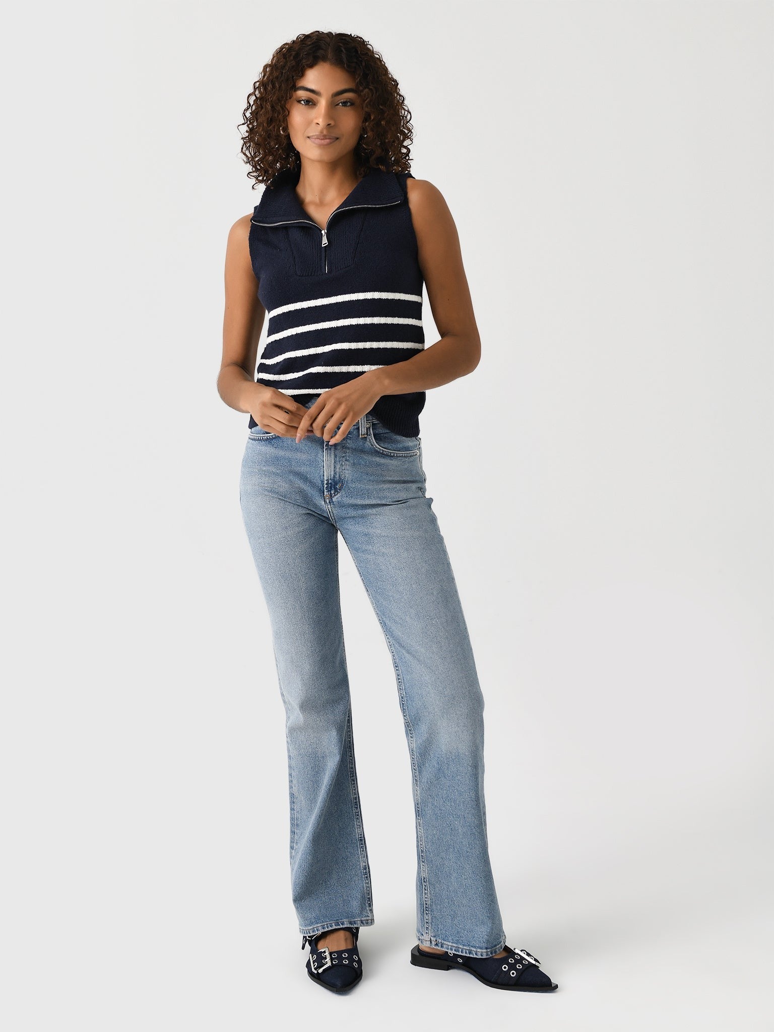 A woman with curly hair wears the Matthew Bruch Quarter Zip Navy Knit Striped Knit Nest, paired with light blue flared jeans and pointed navy flats. She stands against a plain white background in a relaxed pose, holding her hands together near her waist.