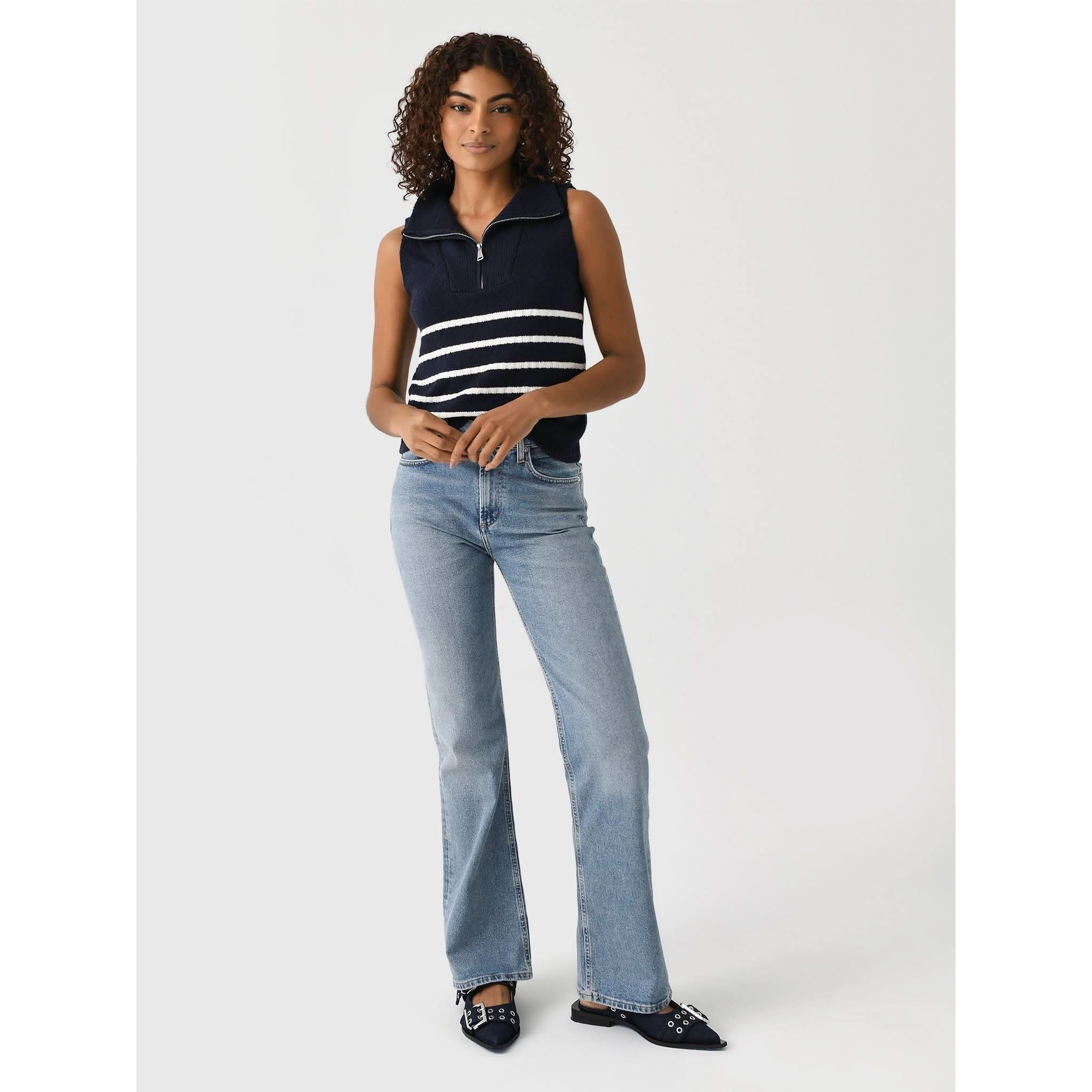 A woman with curly hair wears the Matthew Bruch Quarter Zip Navy Knit Striped Knit Nest, paired with light blue flared jeans and pointed navy flats. She stands against a plain white background in a relaxed pose, holding her hands together near her waist.